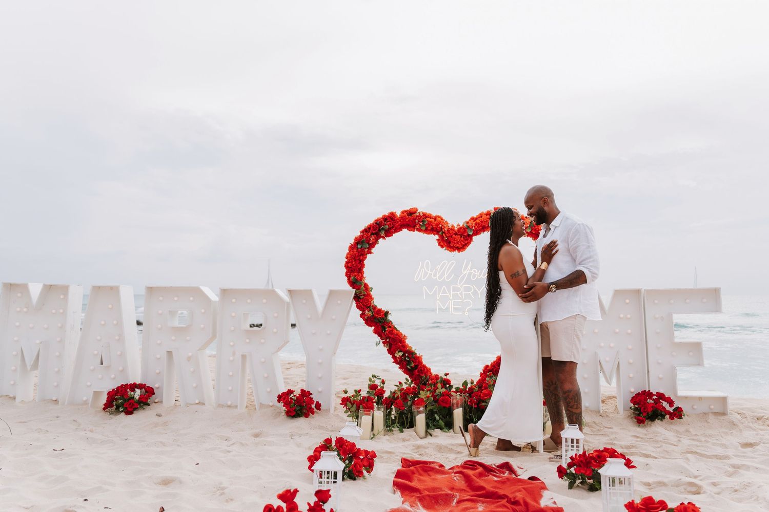 Couple kissing under a red rose heart arch on a beach with MARRY ME signs and rose petals for a romantic proposal.