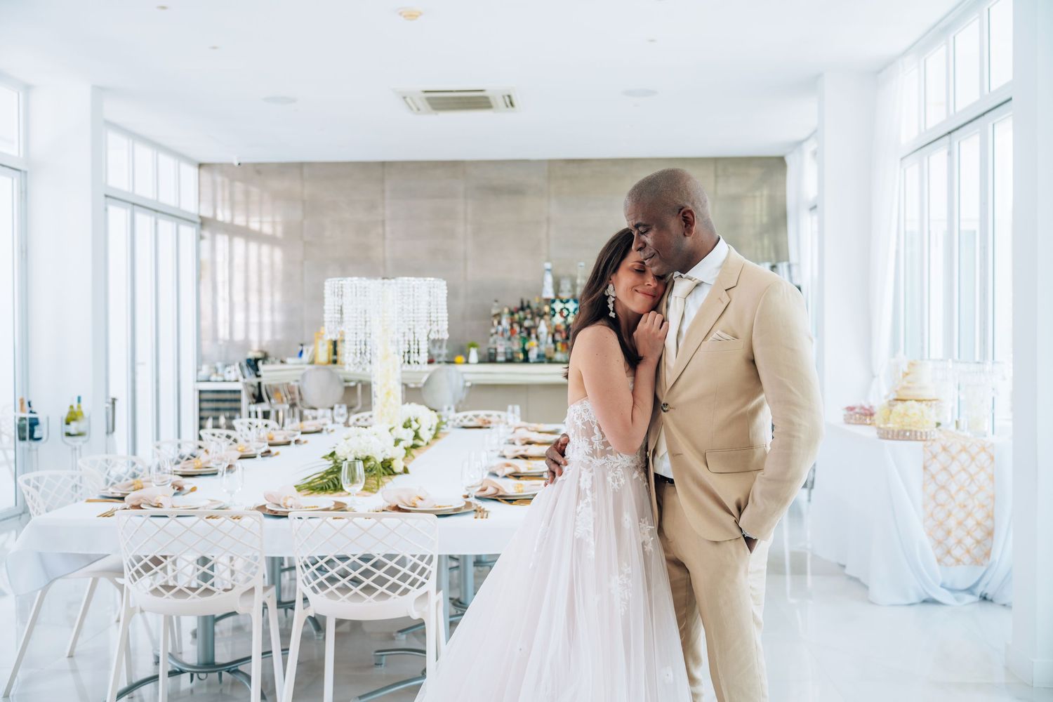 Elegant couple in wedding attire embrace in a bright, white reception venue with decorated tables.