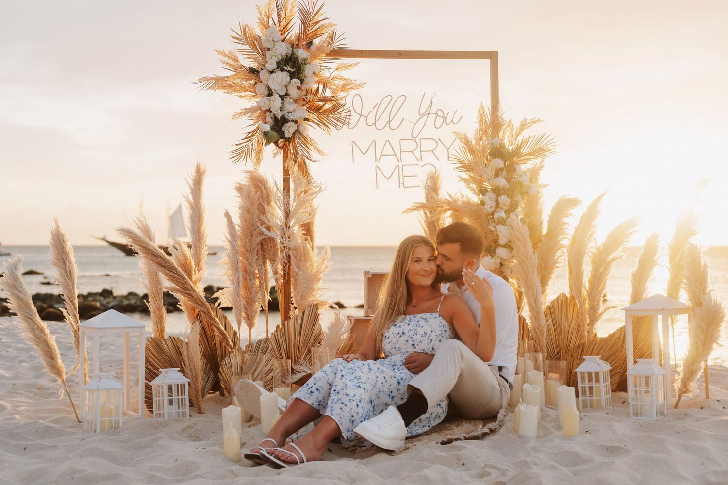 Couple sits together at a romantic beach proposal setup with pampas grass, lanterns, and a Will You Marry Me sign at sunset.