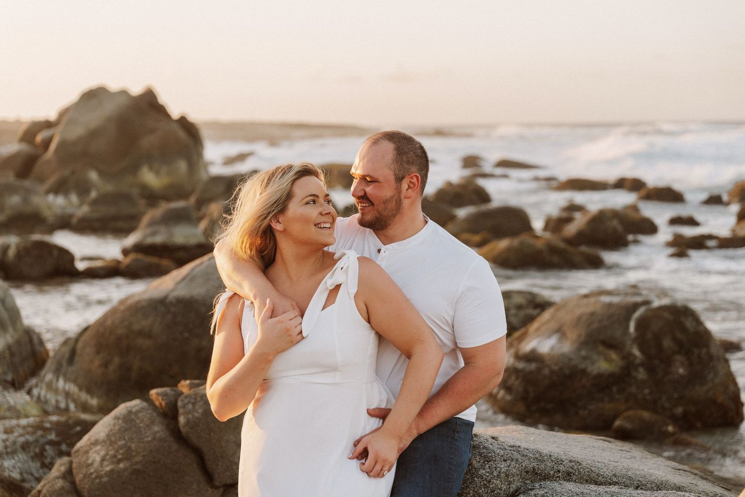 Romantic couple embraces on rocky beach during sunset photoshoot in white outfits.