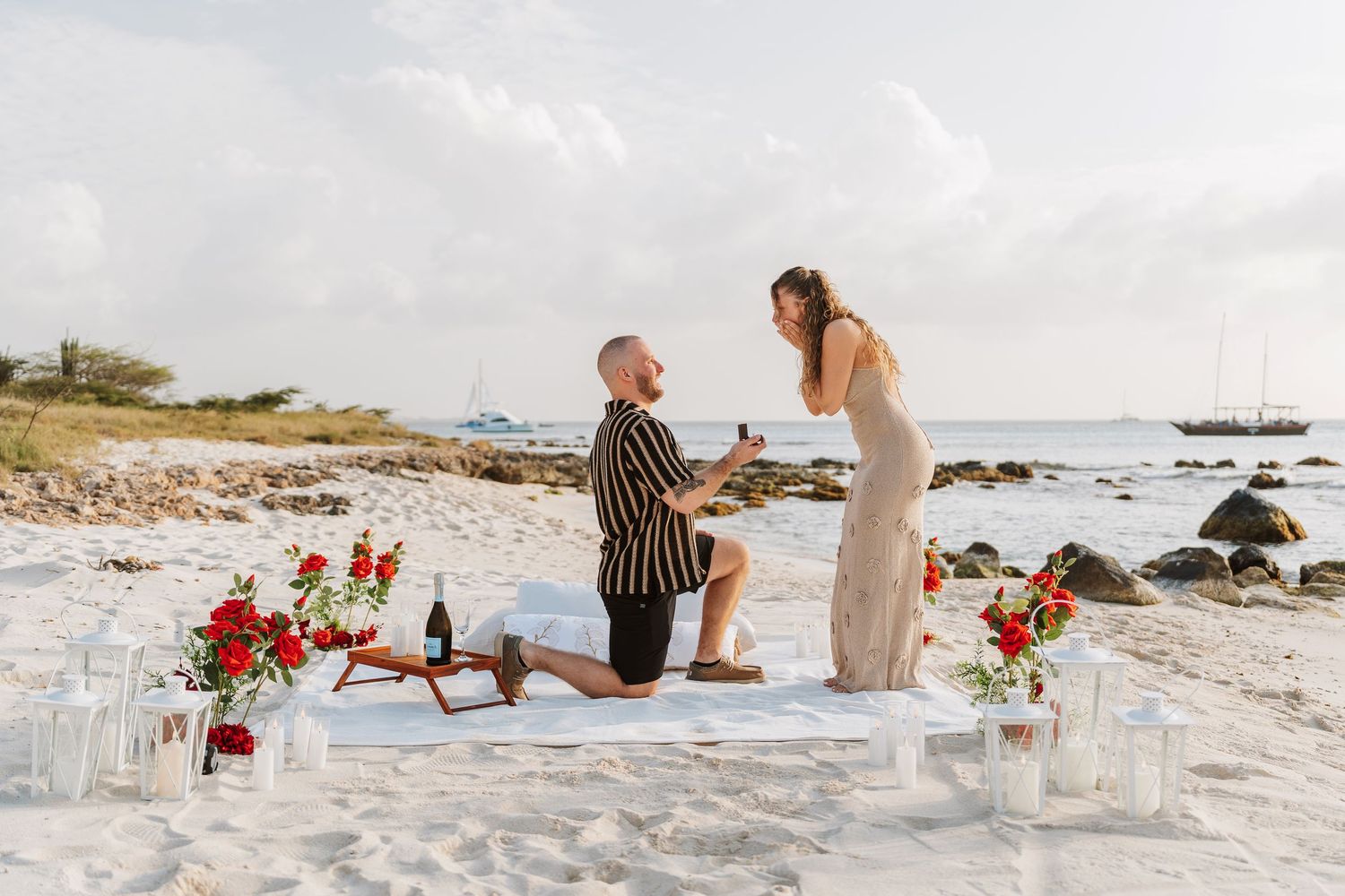 Man proposes to woman on white sand beach with red flowers and candles at sunset.