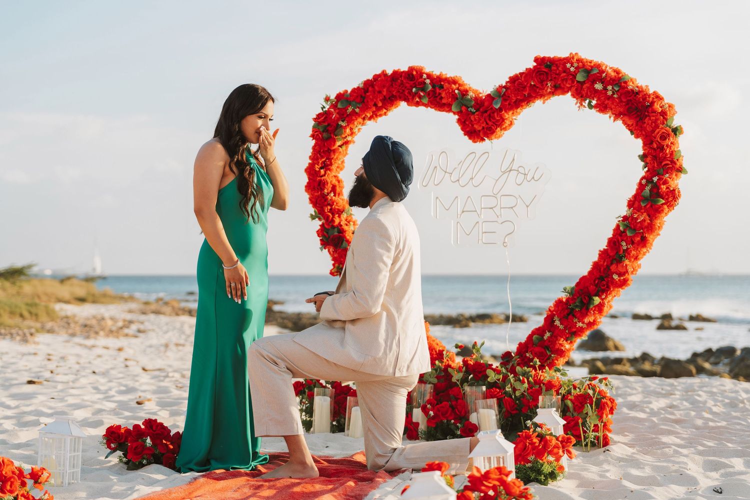 Man proposes to woman on beach with red rose heart arch and romantic floral decorations at sunset.