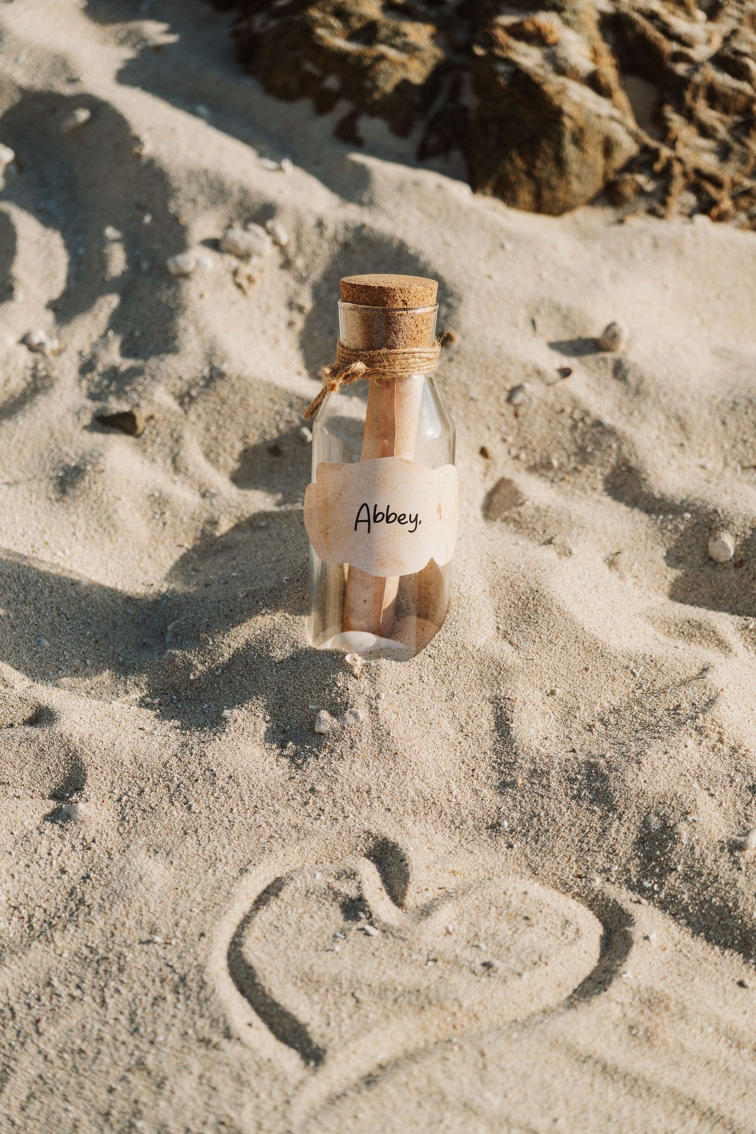A small glass bottle with cork stopper labeled Abbey sits in sandy beach with a heart drawn in the sand nearby.