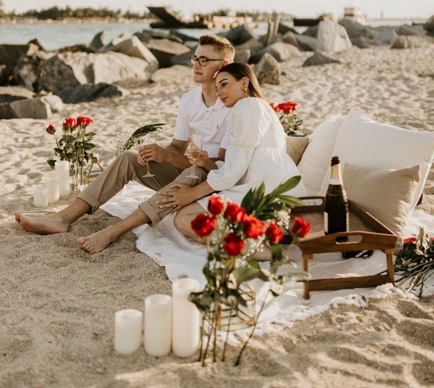 A romantic beach picnic setup with white candles, red roses and a cozy blanket on the sand at sunset.