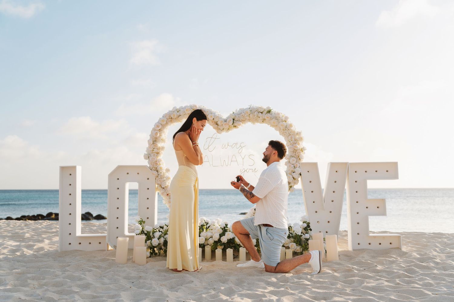 Man proposes to woman on beach with large LOVE letter decorations and floral heart arch by the ocean.