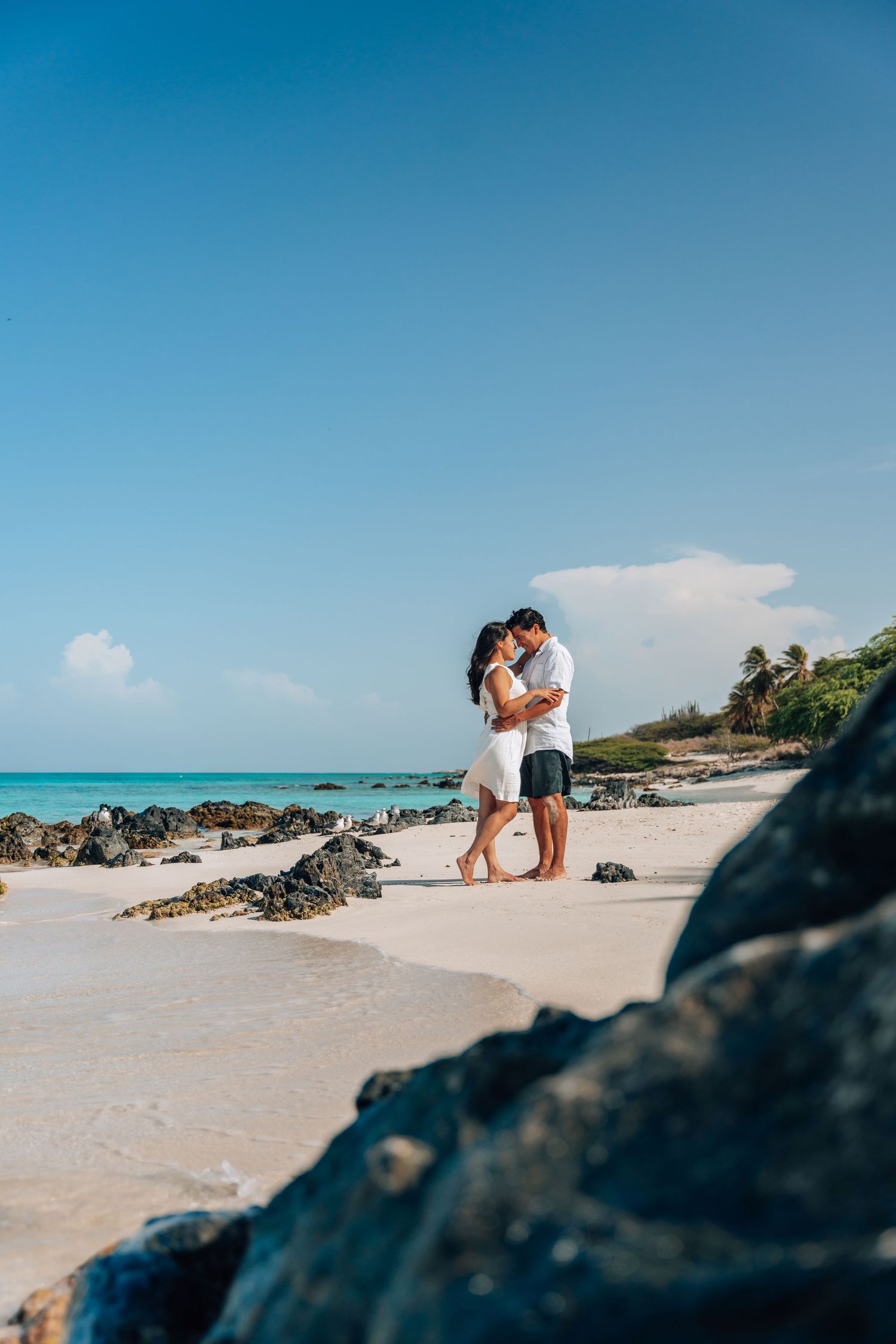 A romantic couple embraces on a tropical beach with turquoise waters and volcanic rocks in Bermuda.