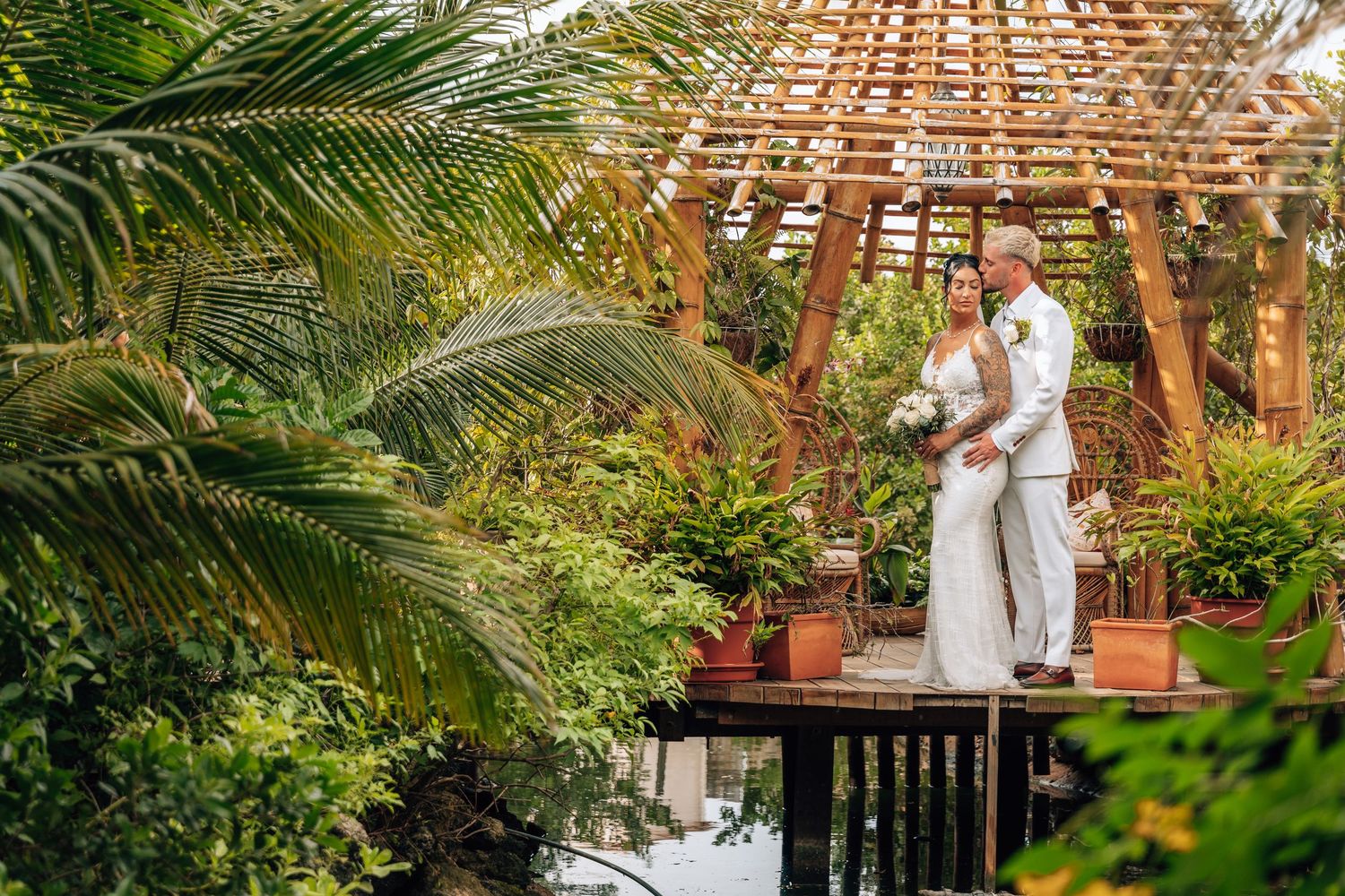 Bride and groom stand under a wooden pergola surrounded by lush tropical greenery on their wedding day.