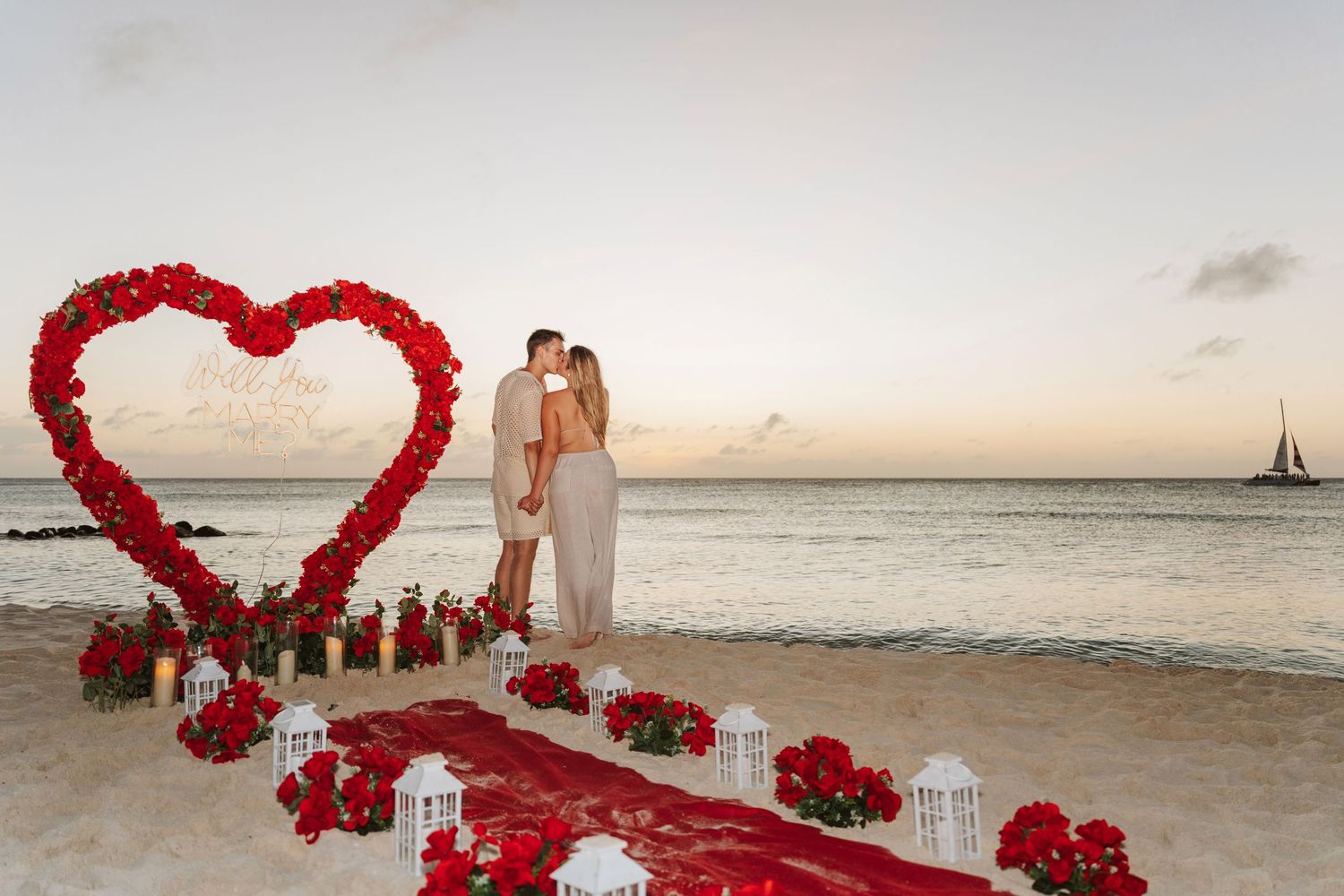 Red heart-shaped floral arch with white chairs on a tropical beach at sunset creates a romantic wedding setting.