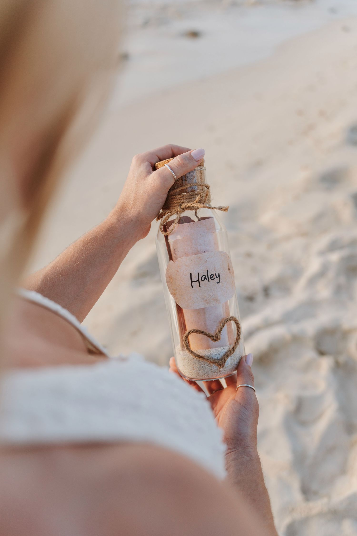 Woman holding a personalized message in a bottle labeled Haley with twine and heart decoration on a sandy beach.