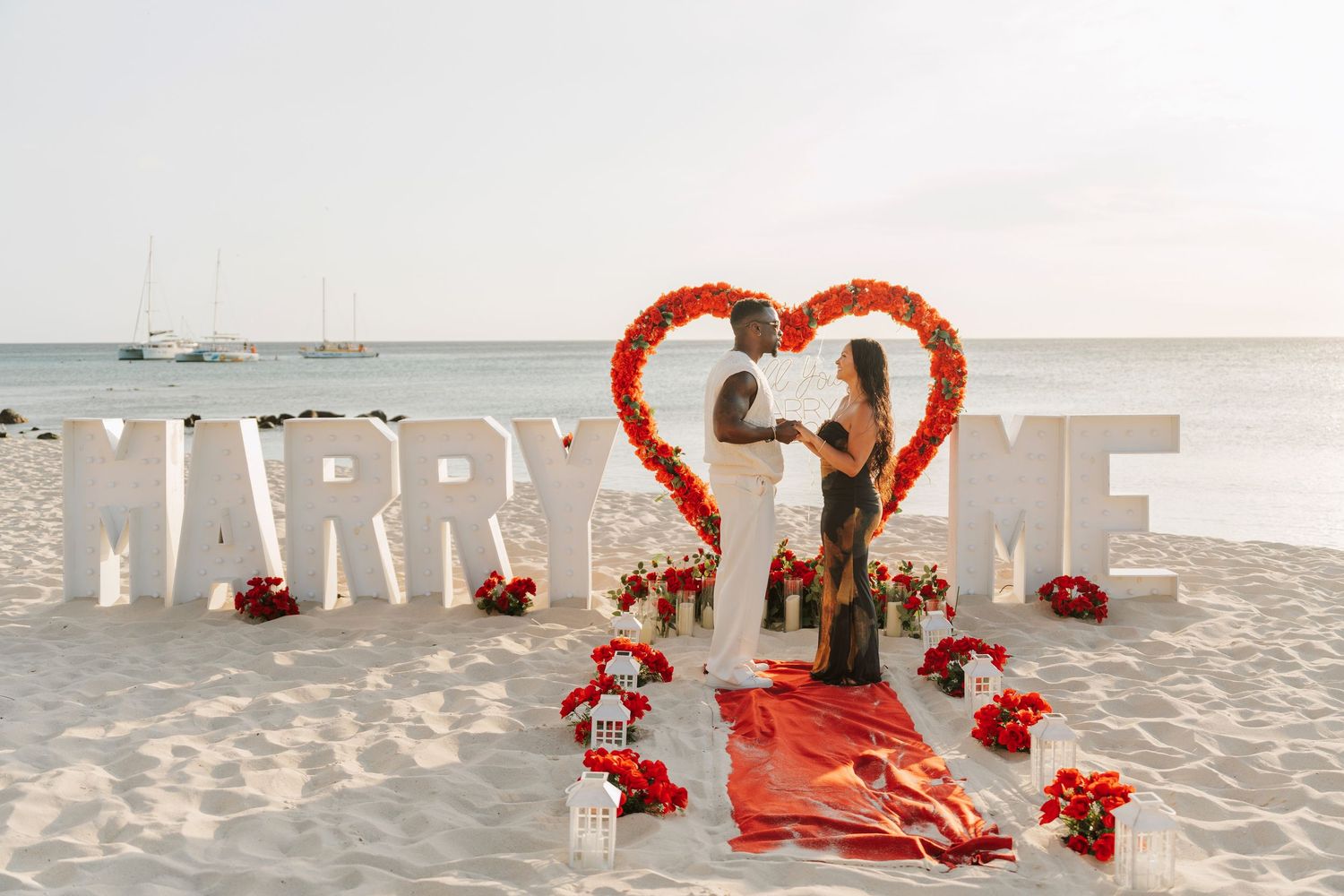 Large white MARRY ME letters with red heart decorations set up on a sandy beach for a romantic proposal.