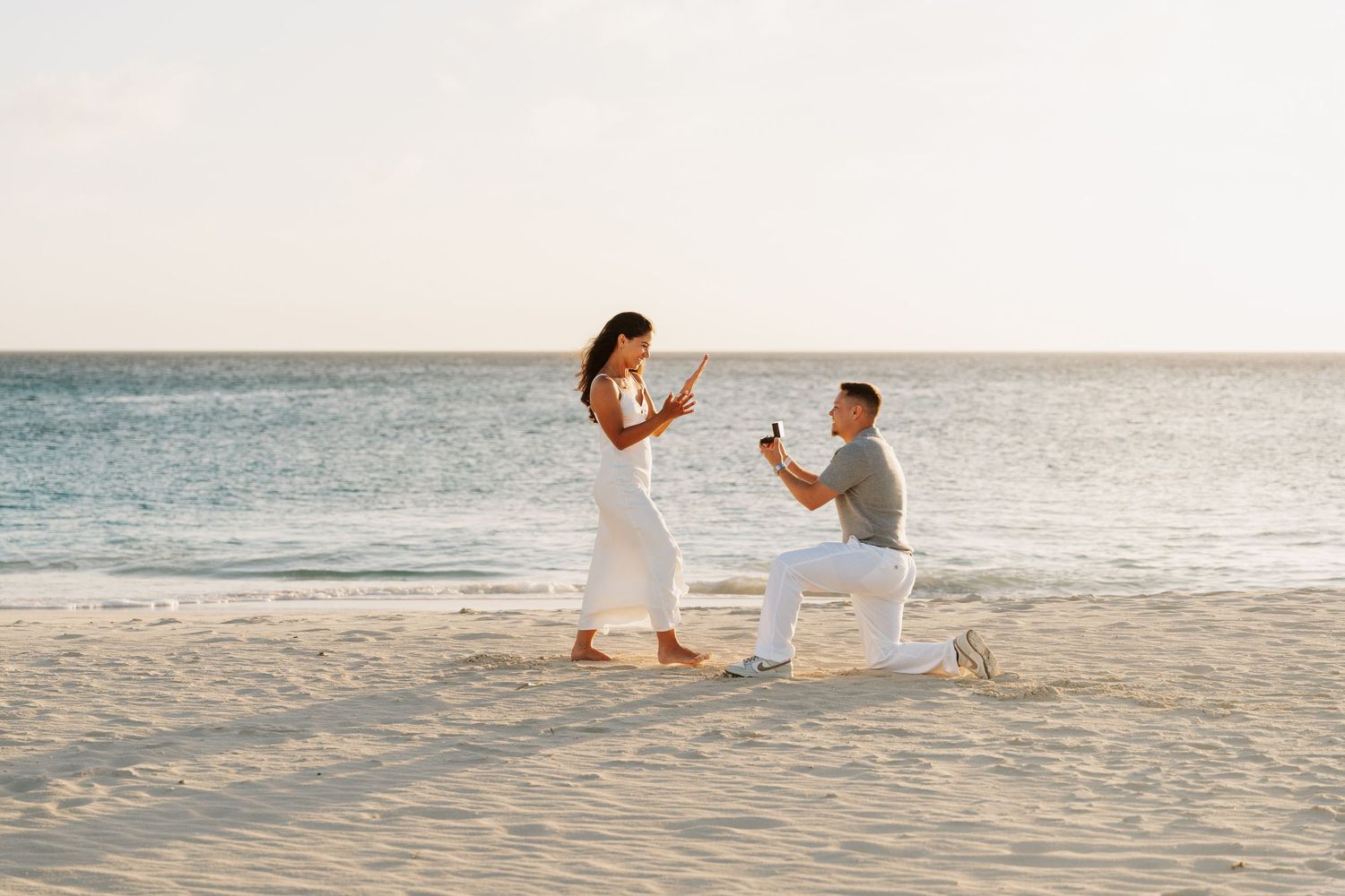 Man proposing to woman on a sandy beach at sunset, kneeling with engagement ring while she reacts with joy.