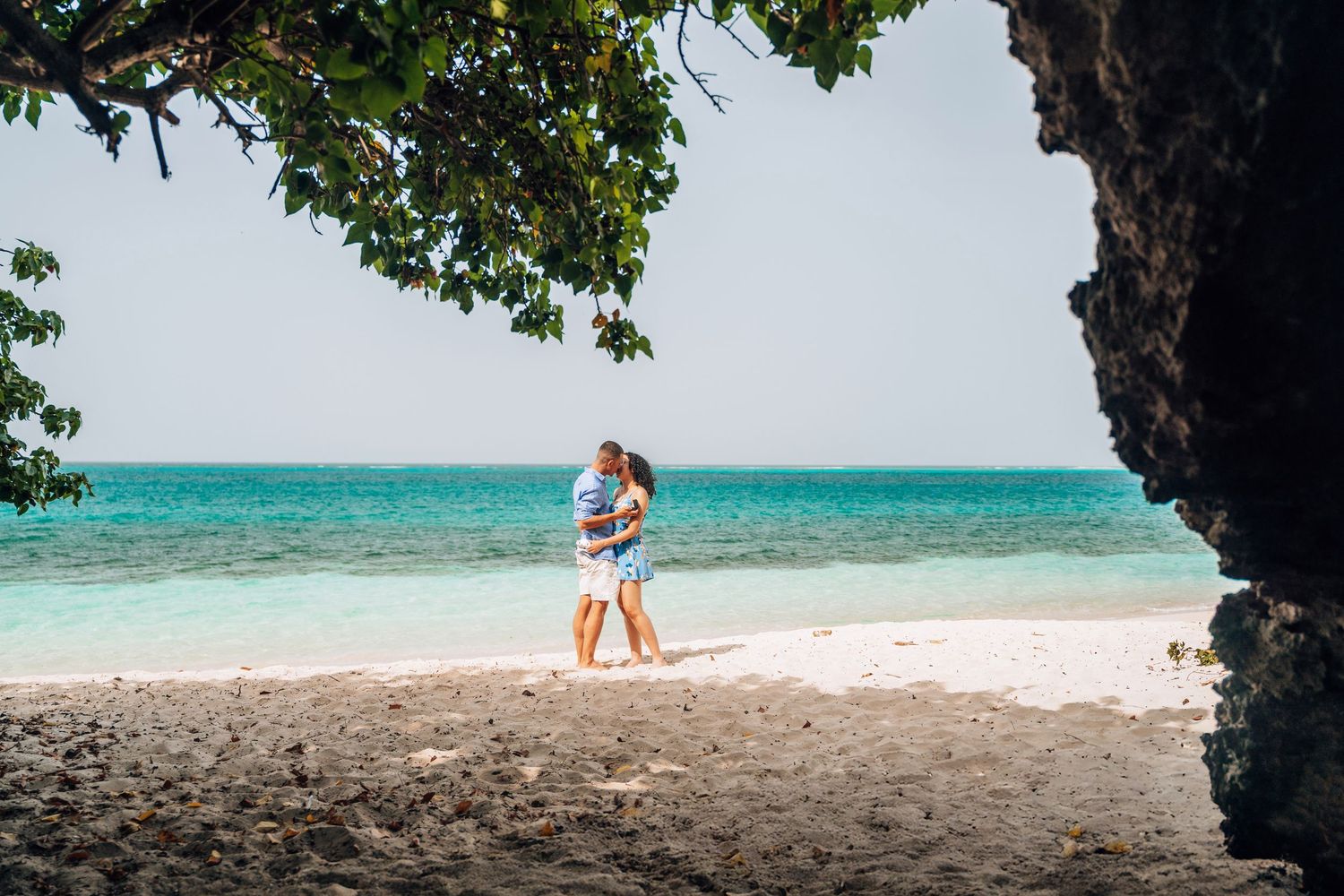 A couple strolls along a pristine white sand beach with crystal clear turquoise waters and lush tropical foliage.