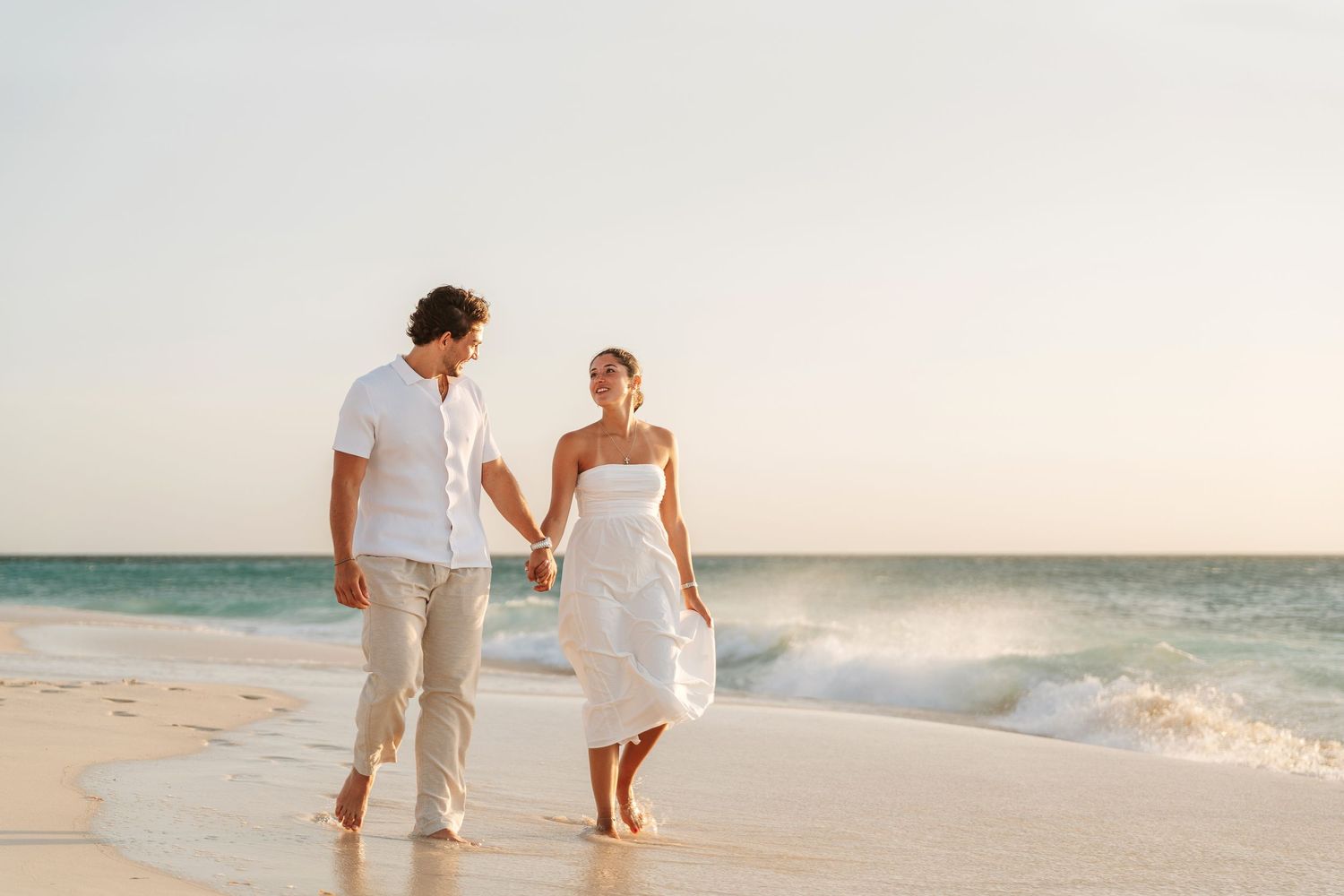 Couple holding hands walking barefoot on a sandy beach at sunset in white outfits with ocean waves in background.