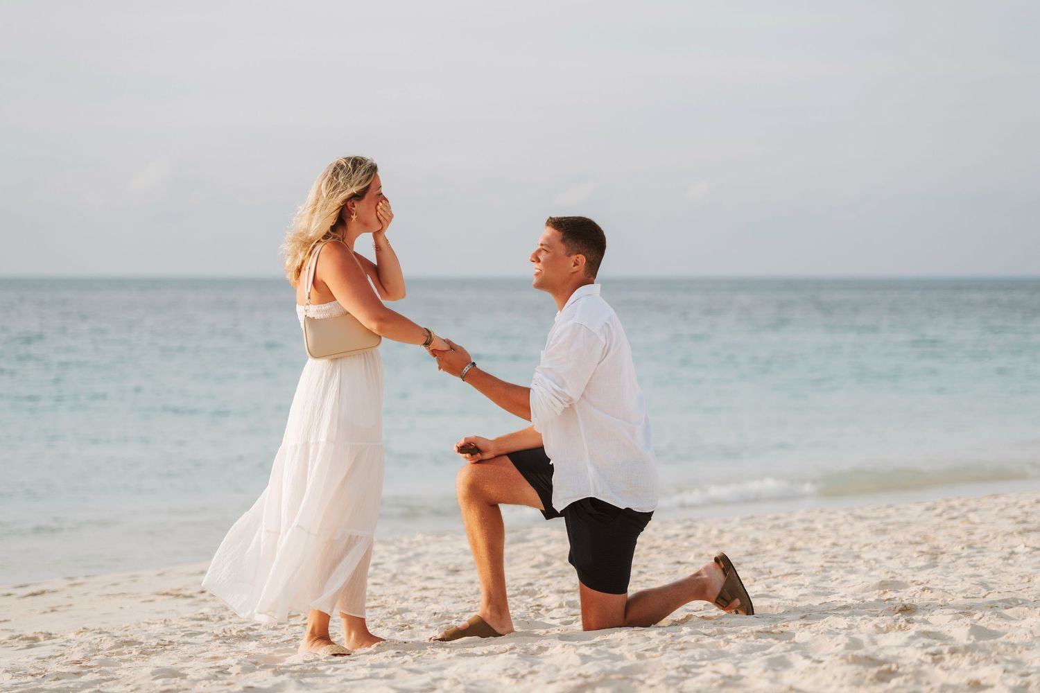 Man proposes to woman on a white sandy beach near the ocean, both dressed in white, sharing a romantic moment.