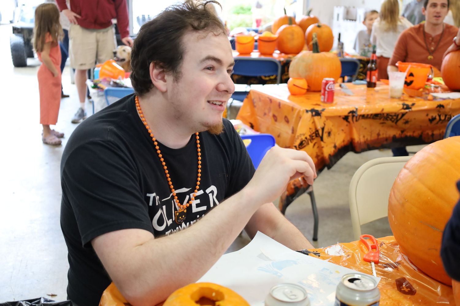 Someone in a black t-shirt carves a pumpkin at a table surrounded by other orange pumpkins during a festive event.