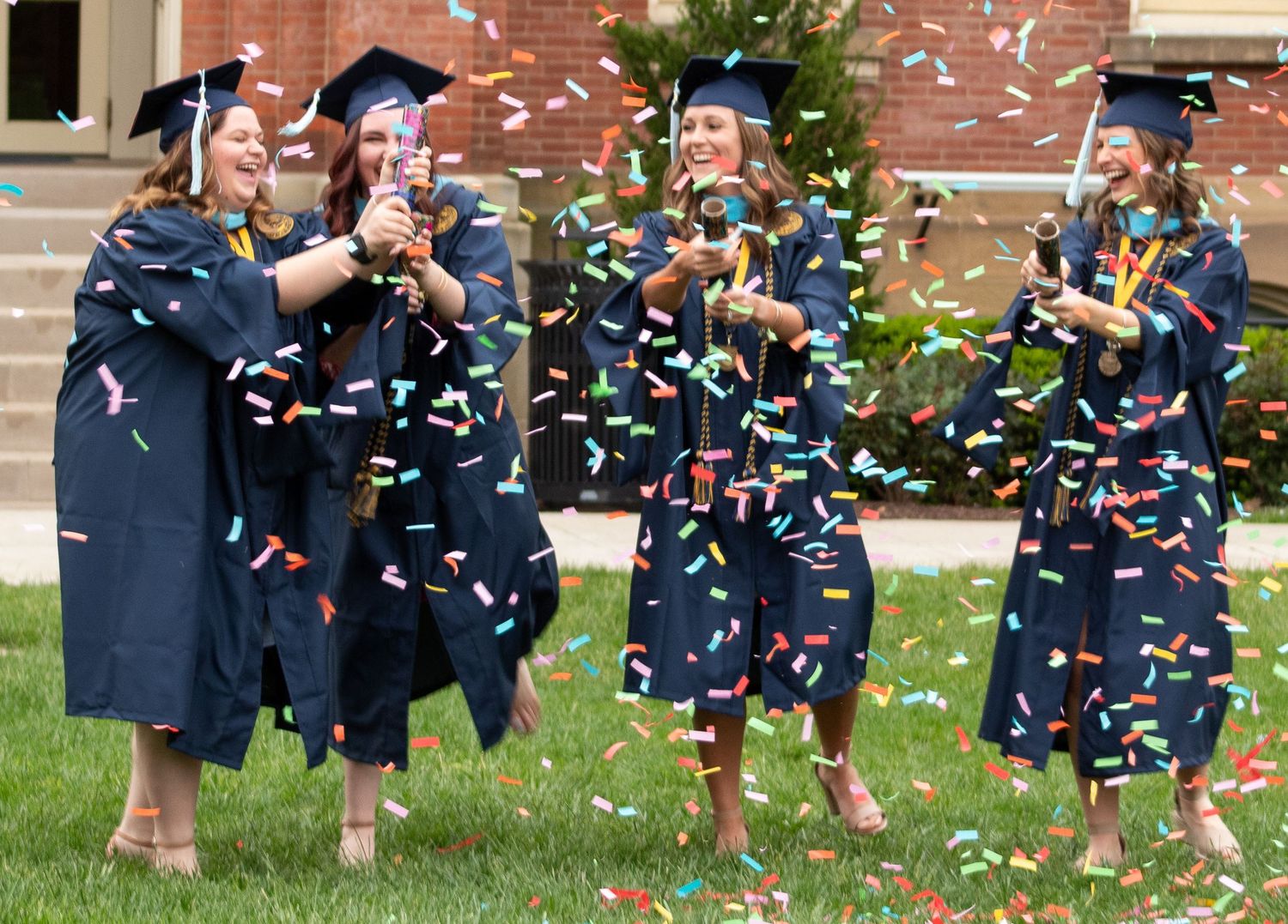 Graduates in black caps and gowns celebrate with confetti during an outdoor commencement ceremony on a grassy lawn.