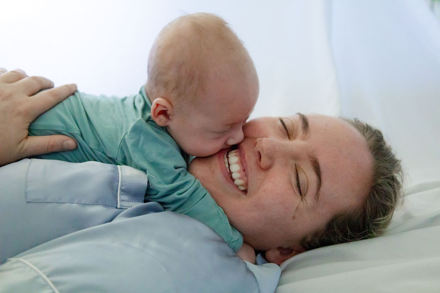 A person lying down in a gray shirt smiles while a baby wearing teal colored clothing leans down toward them.