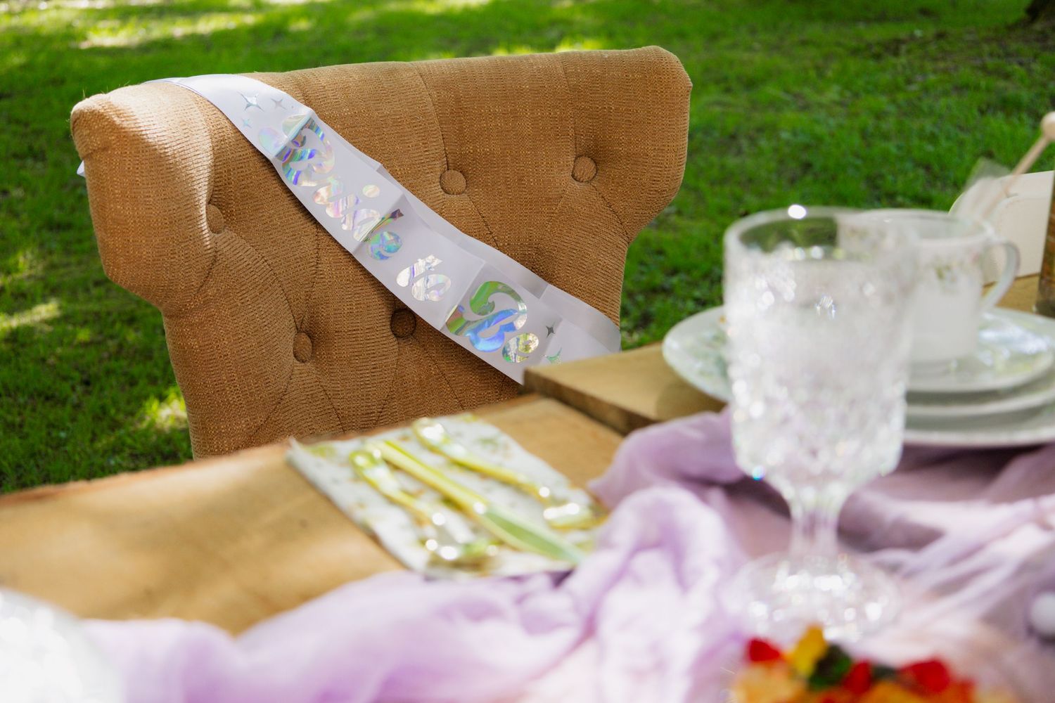 A decorative party sash with iridescent details lies across a brown upholstered chair at an outdoor table setting.