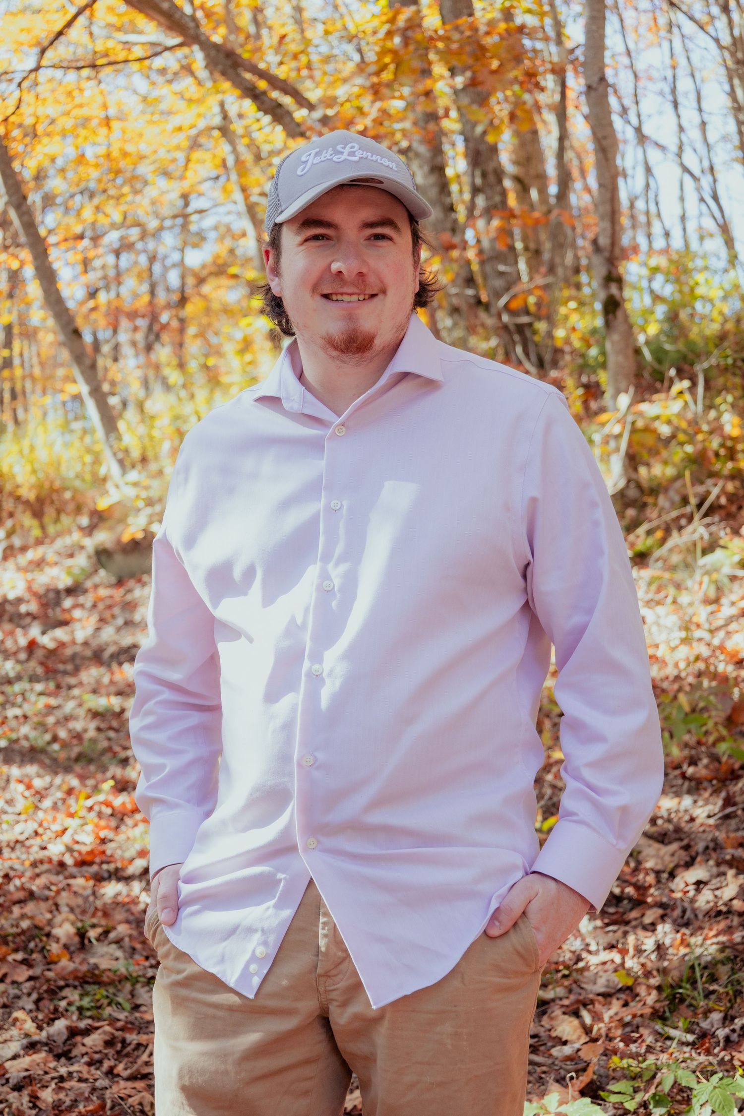 Someone wearing a light pink button-down shirt and khaki pants stands smiling in a colorful autumn forest setting.
