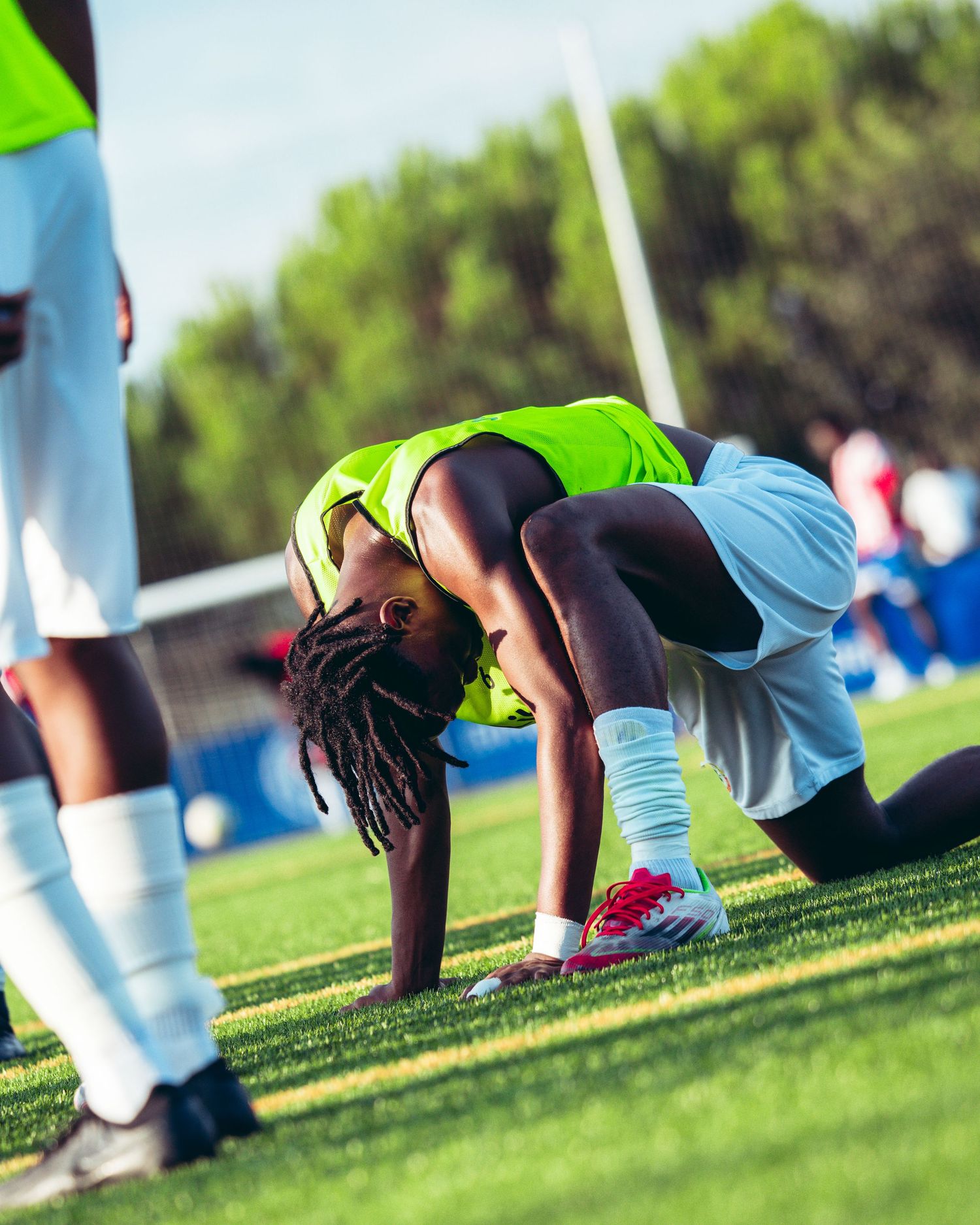 Soccer player in green vest crouches on grass field during a sunny outdoor football match.