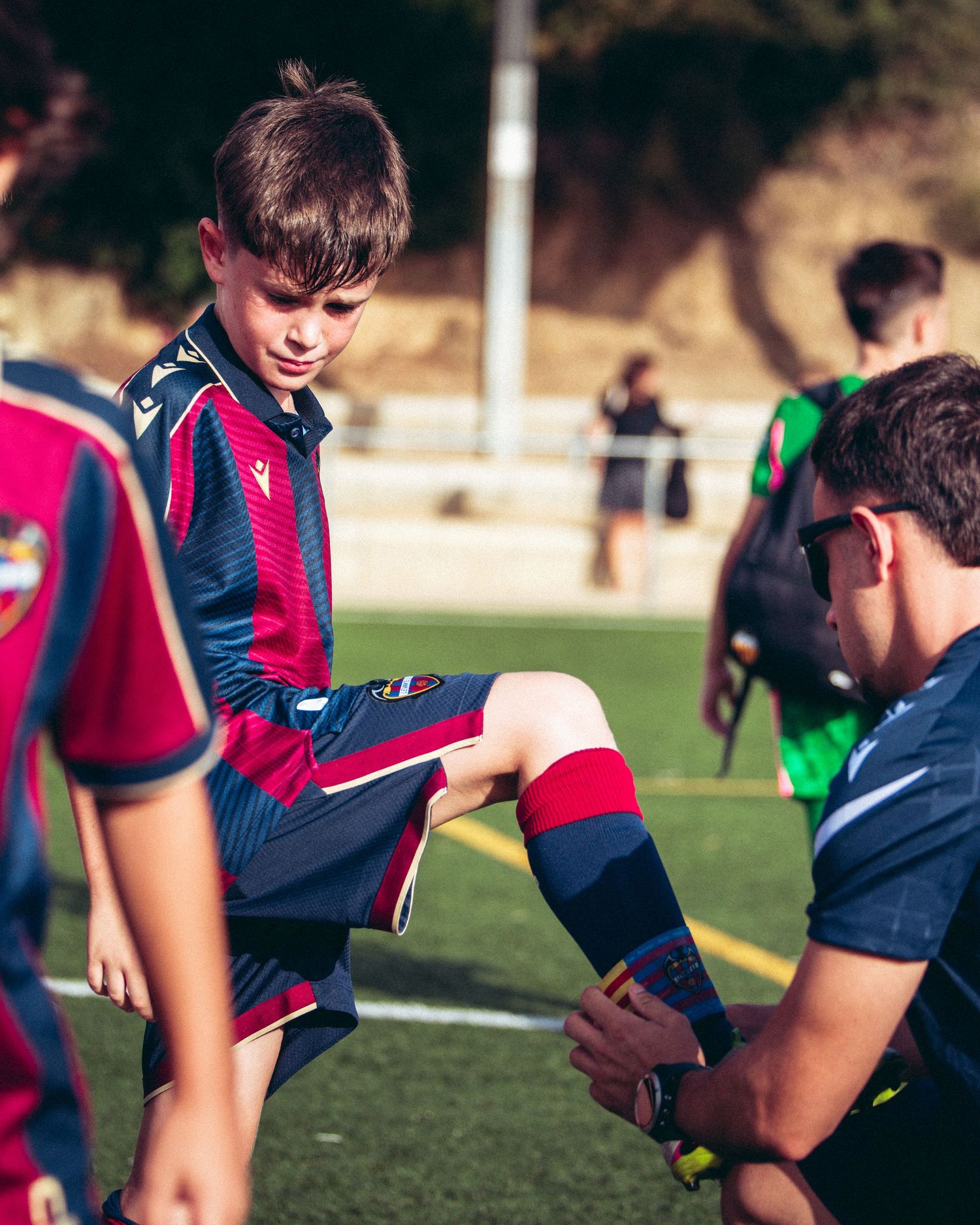 Youth soccer coach adjusting shin guards on a young player in a navy and red striped jersey.