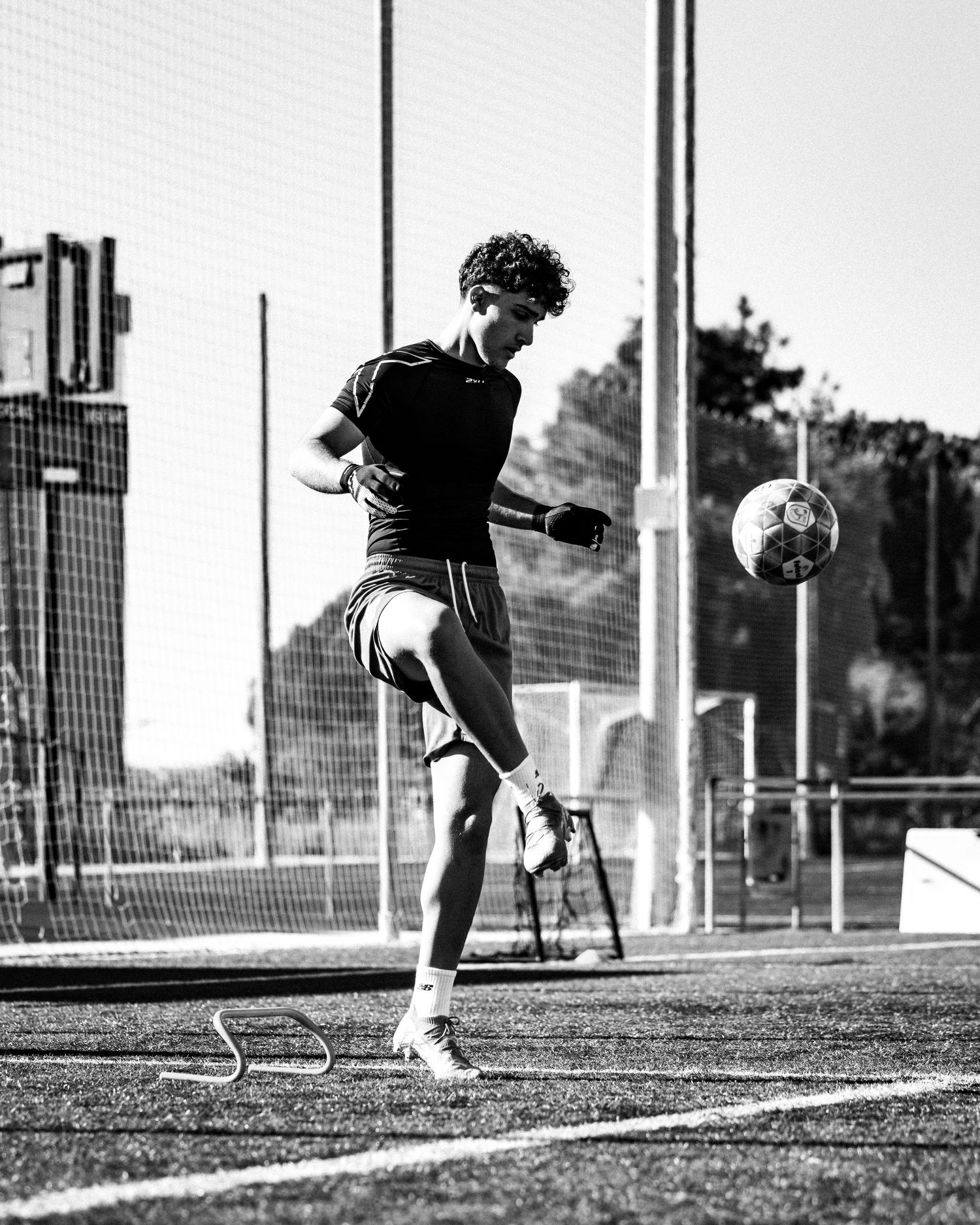 Young soccer player juggling a ball mid-air on an outdoor field in black and white.