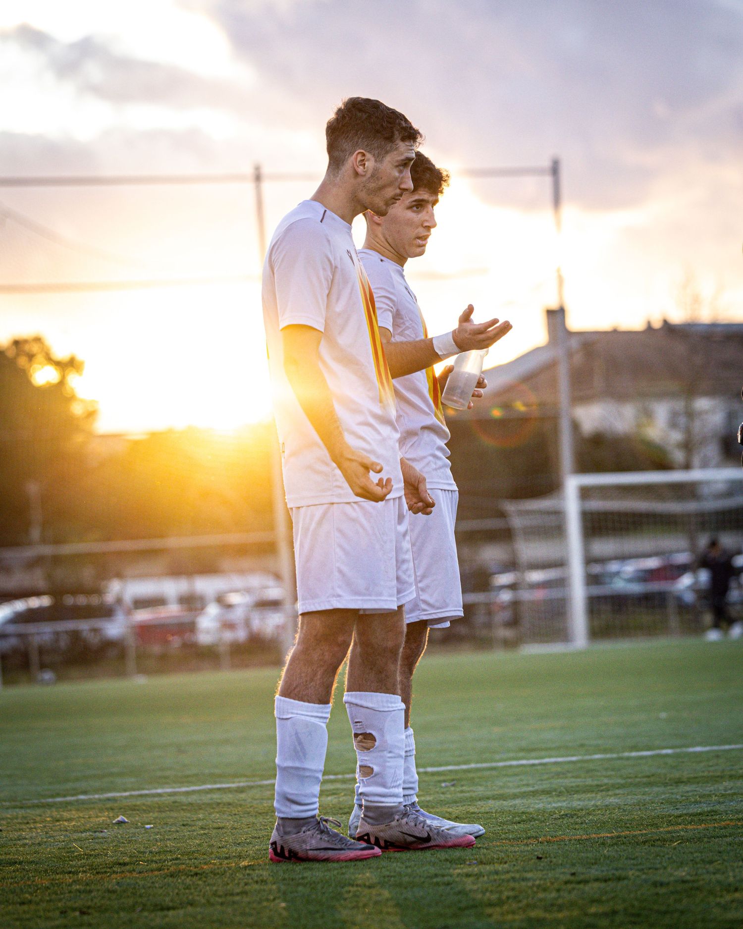 Two young soccer players in white uniforms standing together on field during golden sunset.