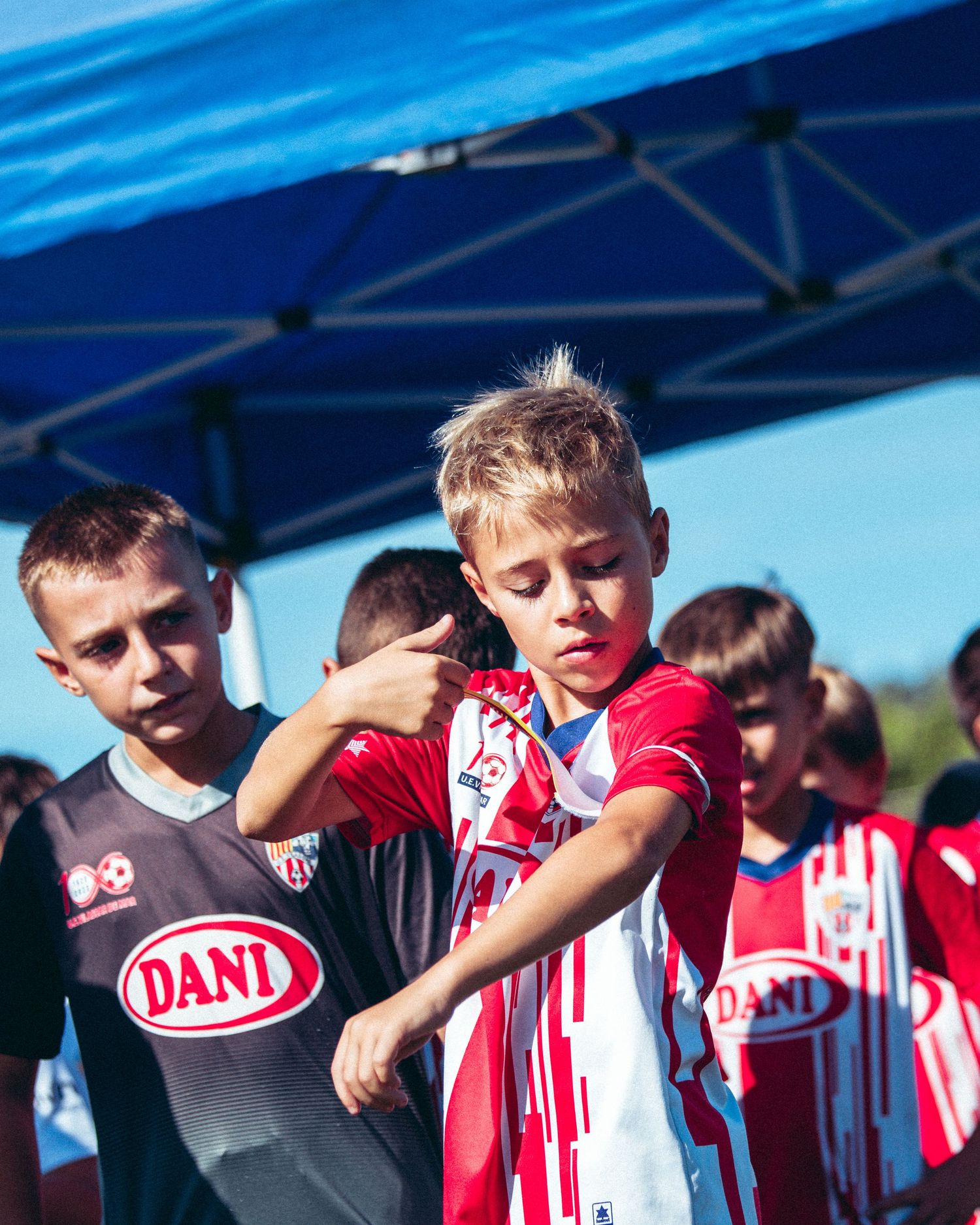 Young boys in soccer uniforms at outdoor tournament under blue canopy on sunny day.