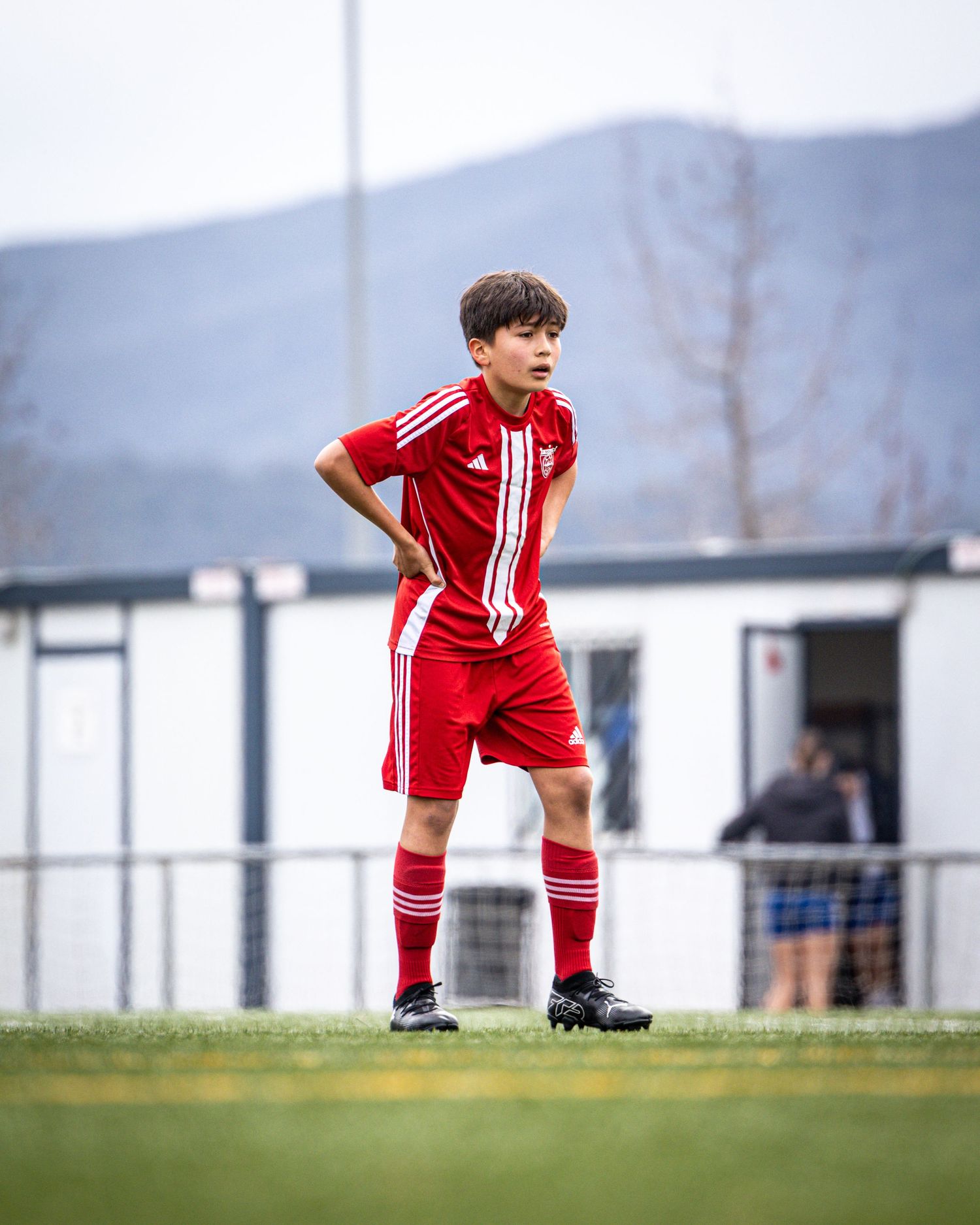 Young boy in red Adidas soccer kit standing on green pitch with mountains in background.