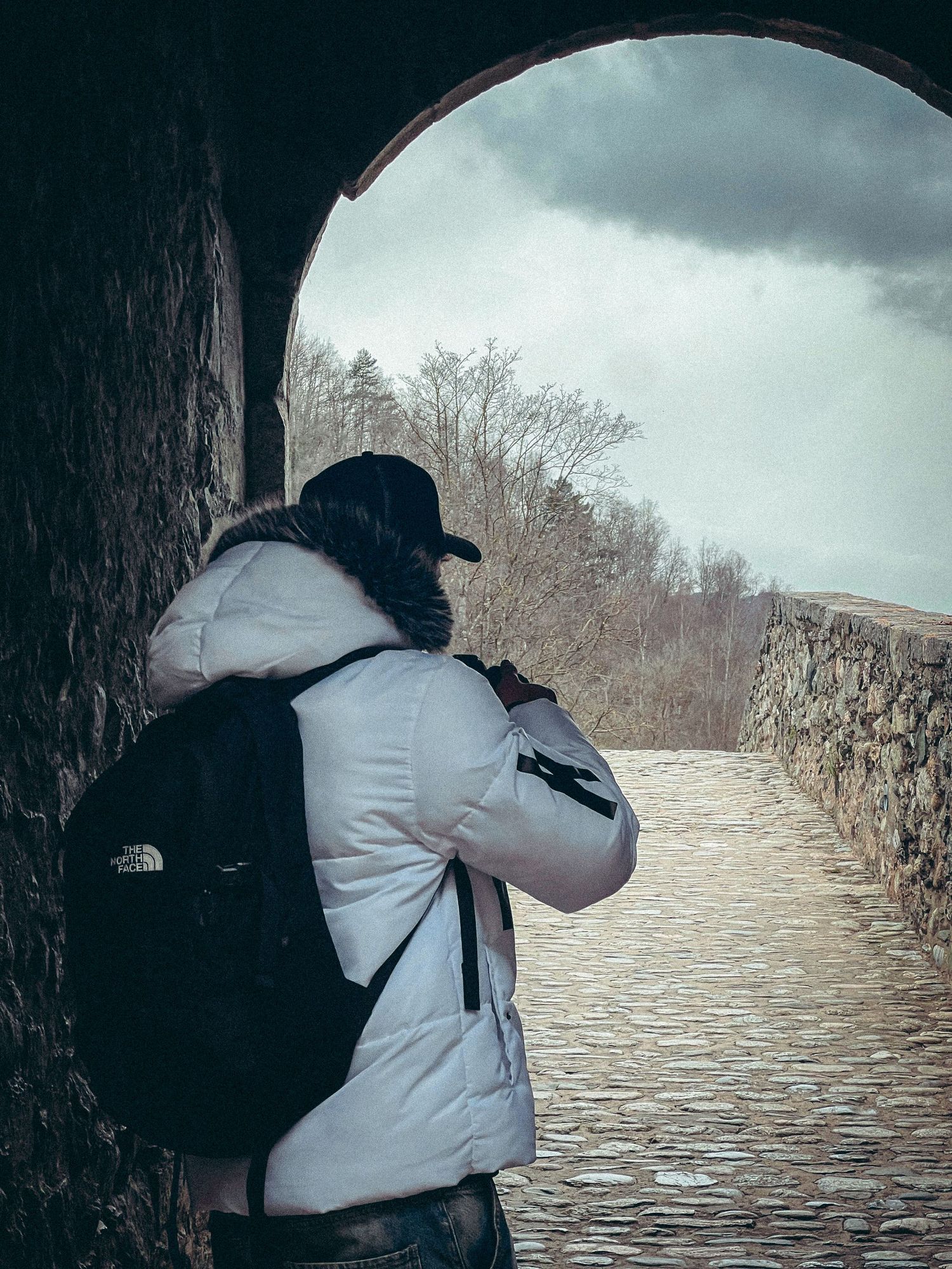 Person in white North Face jacket with backpack standing under stone arch on cobblestone path.