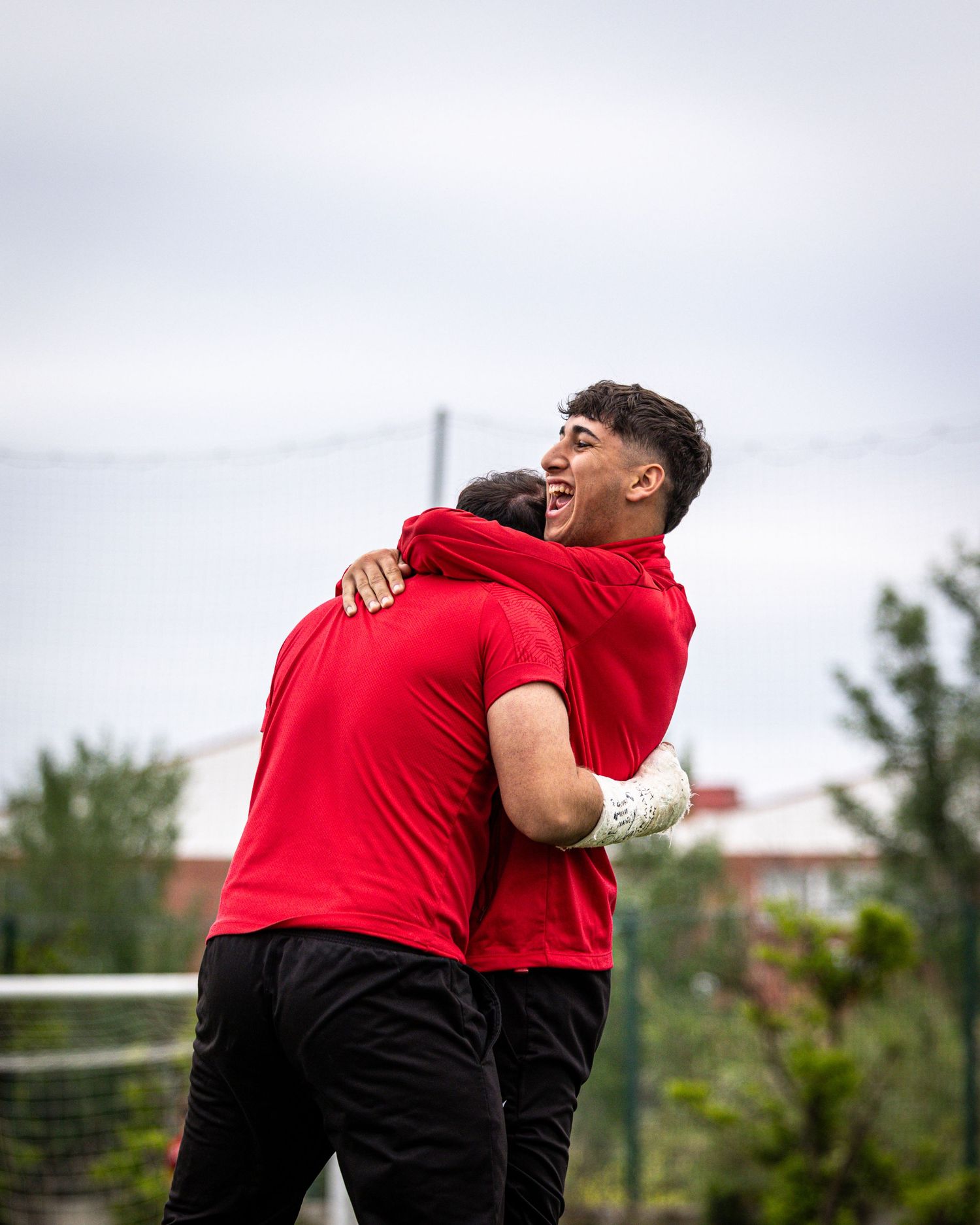 Two soccer players in red jerseys celebrate with a joyful hug on an outdoor football field.