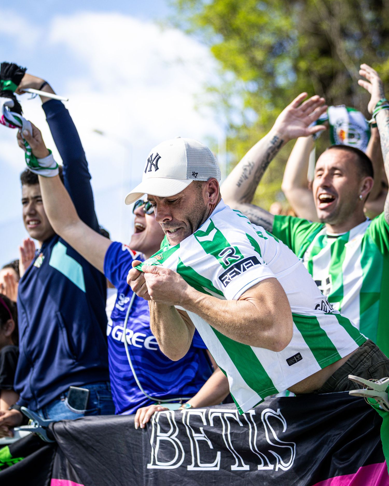 Enthusiastic Real Betis fans cheer and celebrate while holding a Betis banner outdoors.