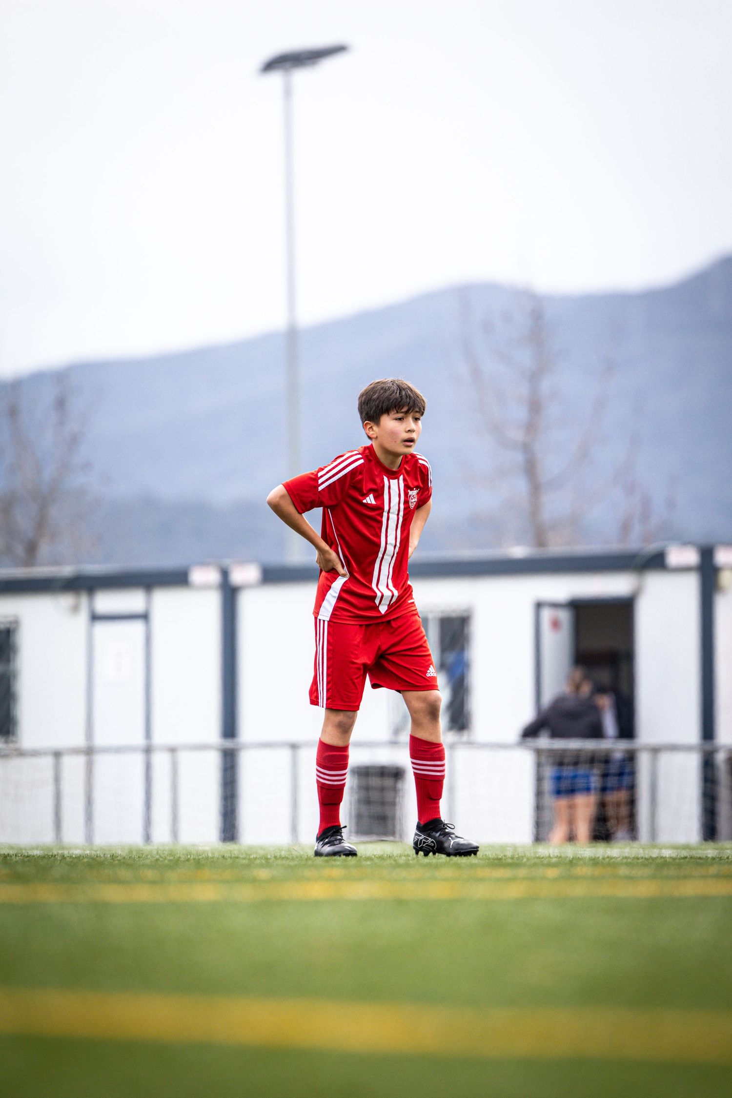 Young boy in red Adidas soccer kit standing on green pitch with mountains in background.