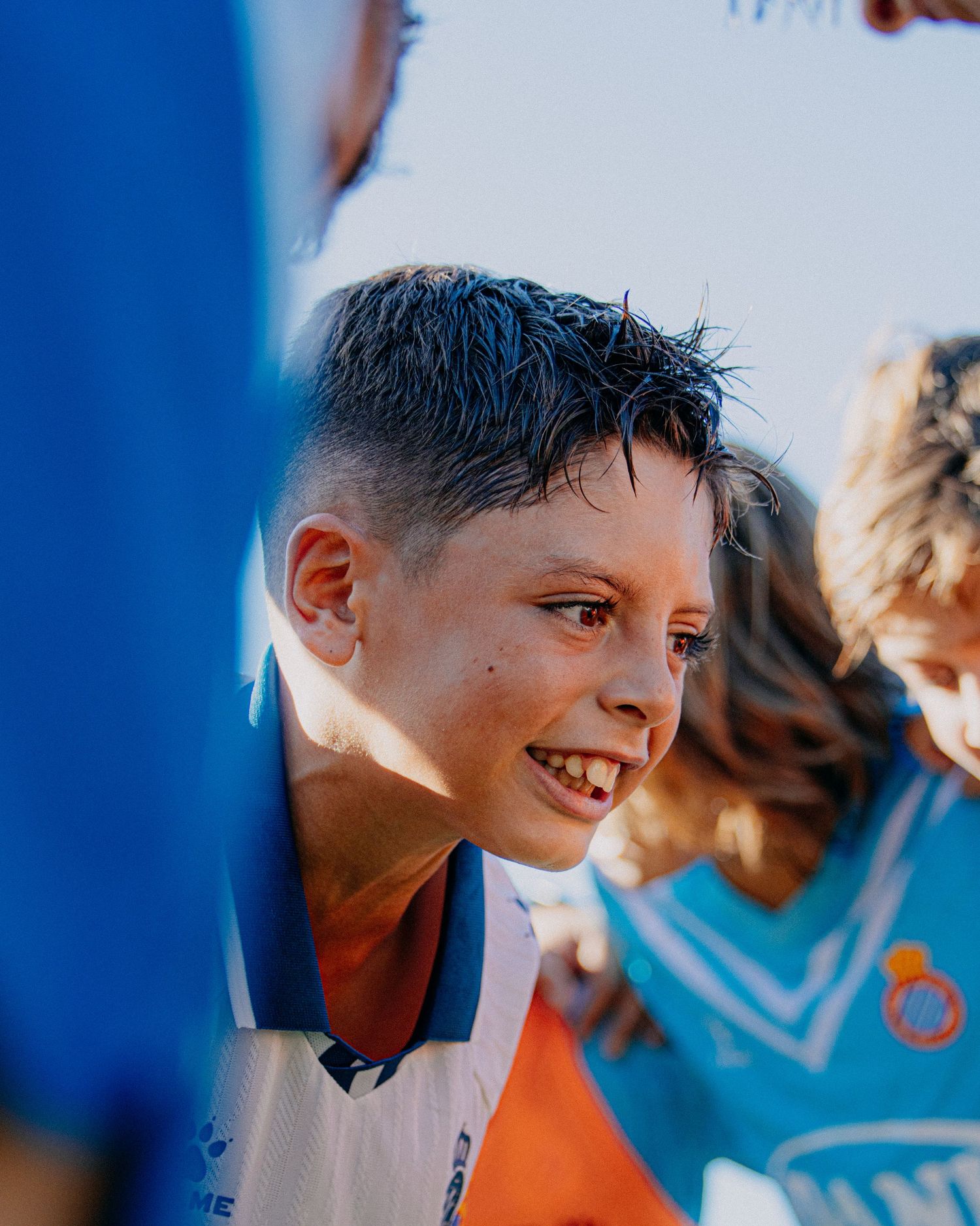 Smiling young boy in a white and orange soccer jersey celebrating with teammates after a match.