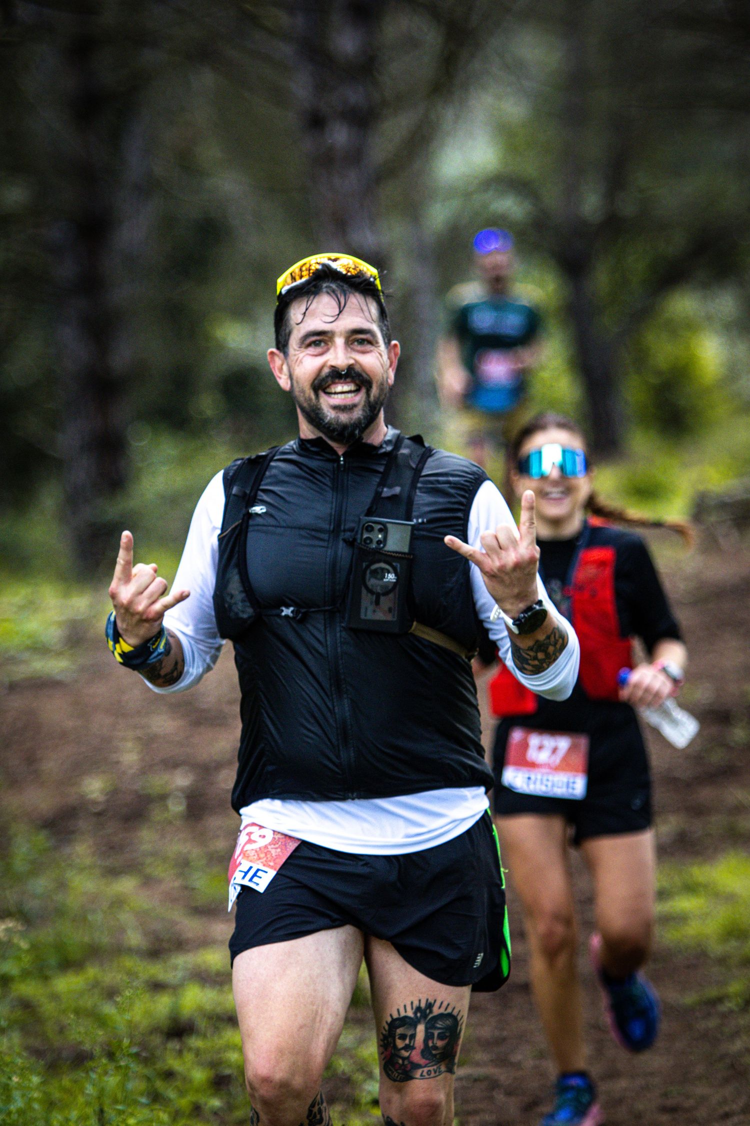 Smiling trail runner makes shaka gesture while competing in forest ultramarathon race with other runners behind.