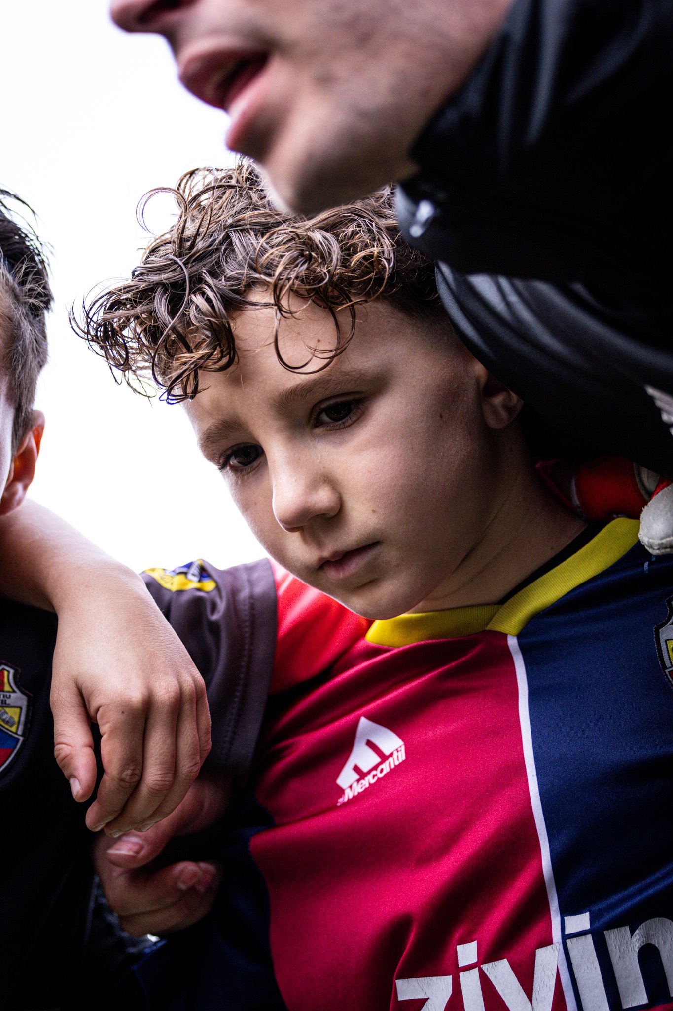 Young curly-haired child in red and blue soccer jersey surrounded by teammates in a huddle.