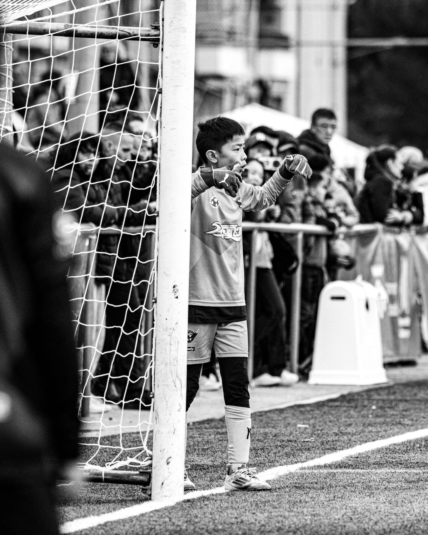 Black and white photo of young goalkeeper standing by goal post during youth soccer tournament match.
