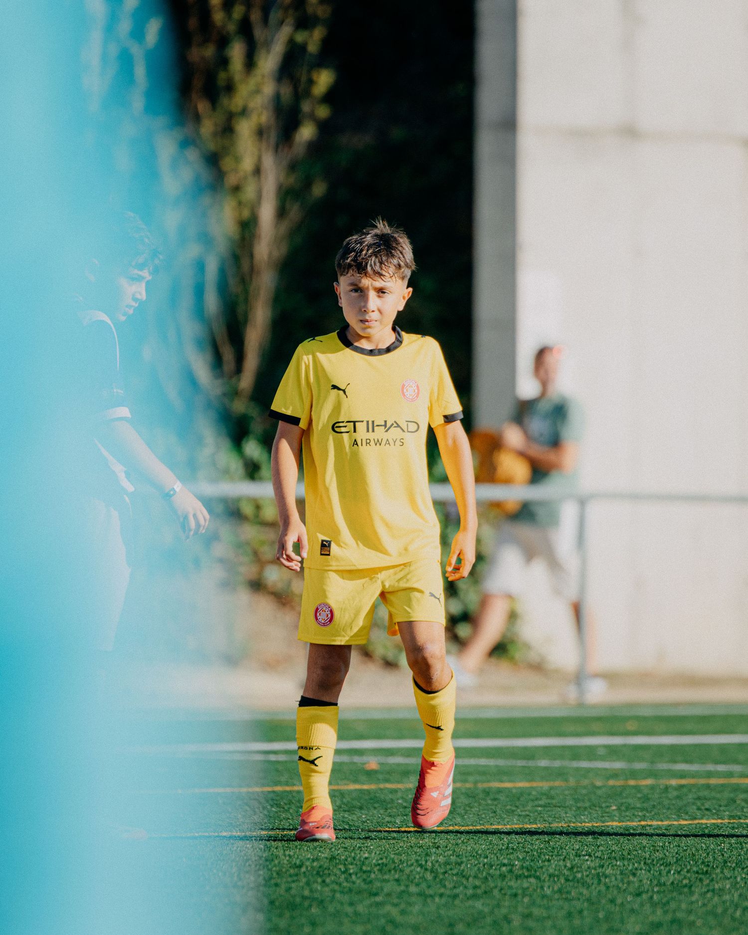 Young boy in yellow Etihad Airways Puma kit walks on a green soccer field.