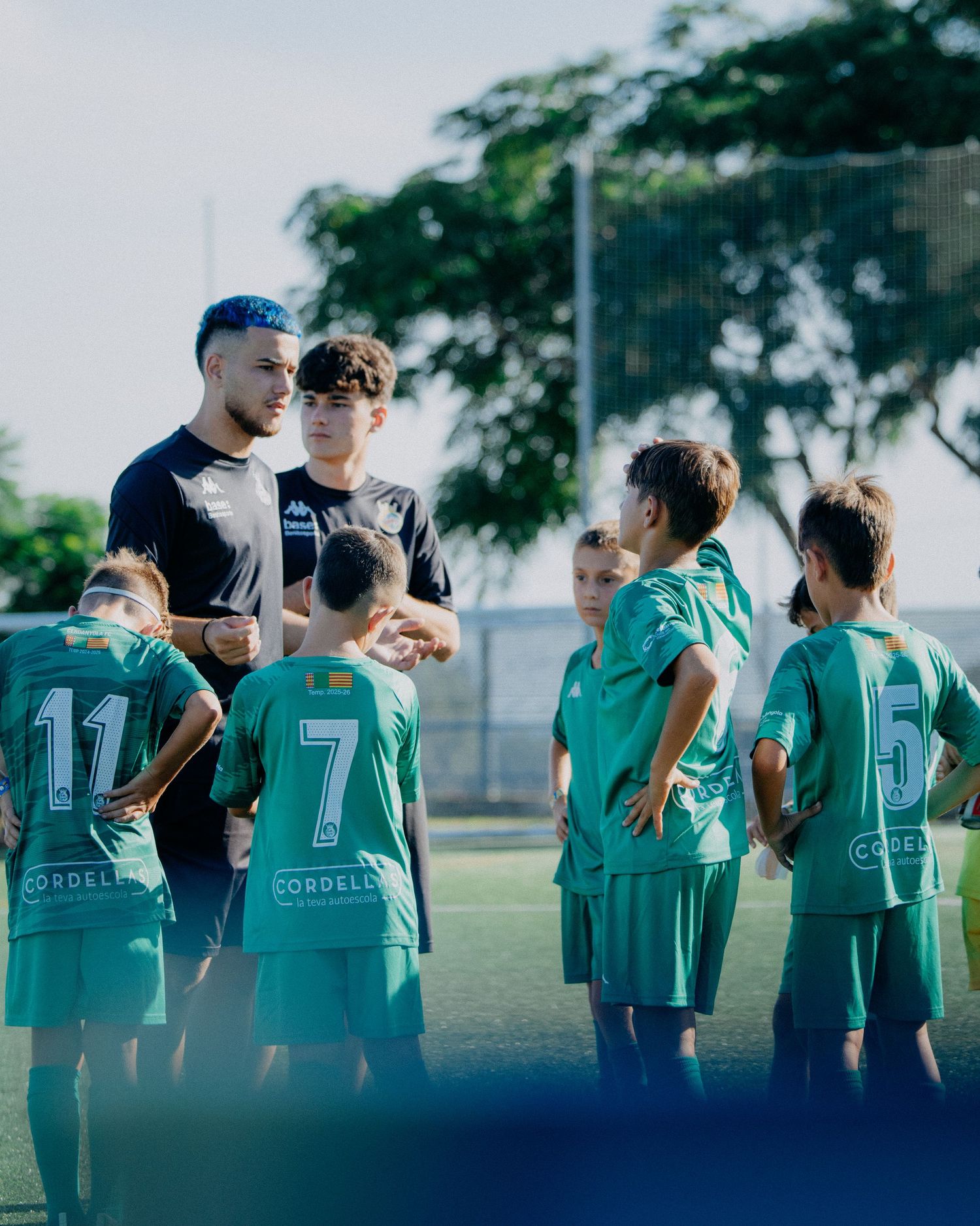 Coach talks to youth soccer players in teal uniforms on a sunny field.