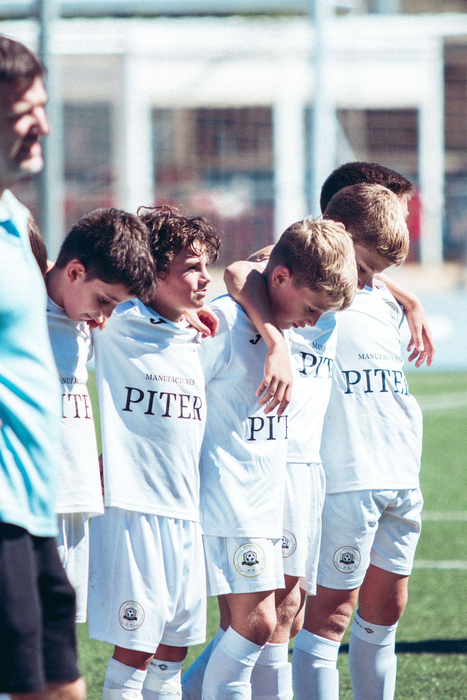 Youth soccer team in white Piter jerseys huddling together on sunny football pitch.