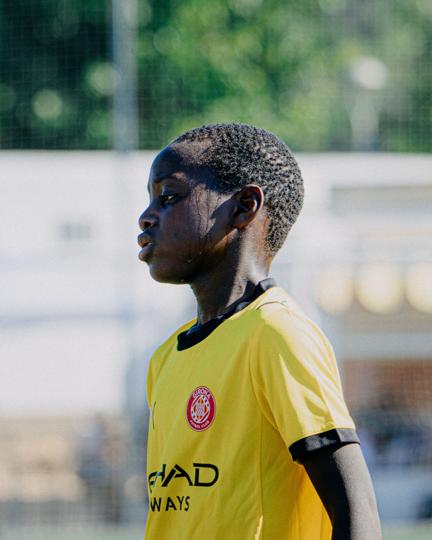Young soccer player in yellow Girona FC jersey looks focused on the field during a sunny outdoor match.