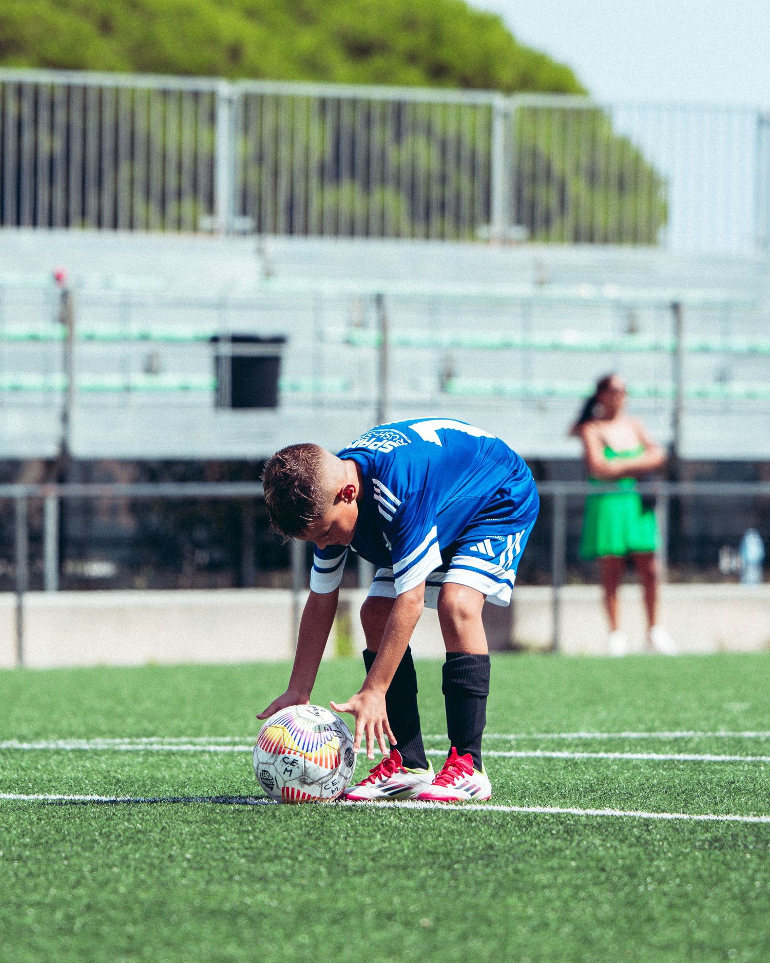 Young boy in blue Adidas soccer kit placing the ball for a free kick on a sunny turf field.