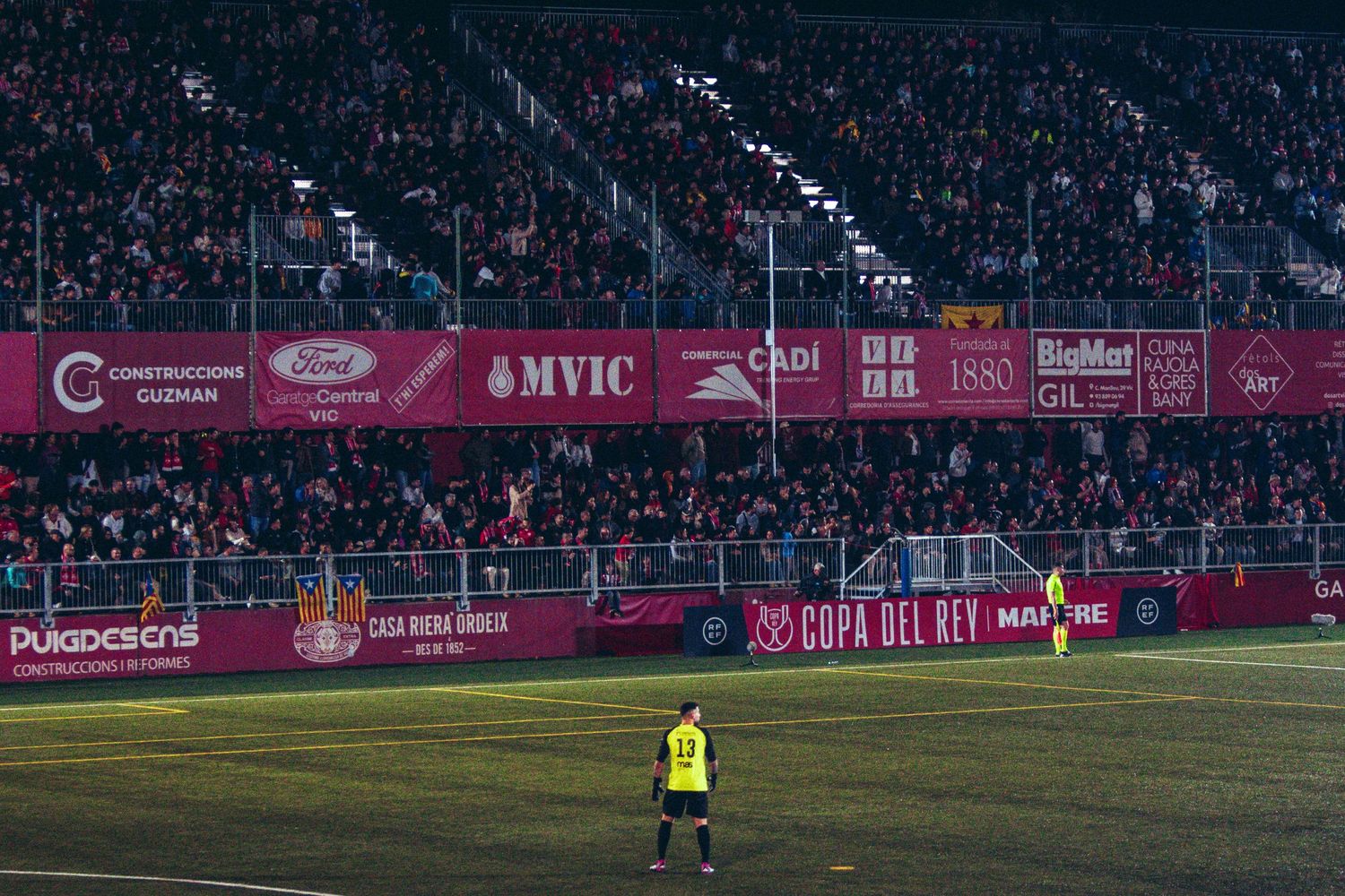 Referee stands on soccer field during Copa del Rey match with packed stadium crowd and sponsor banners in background.