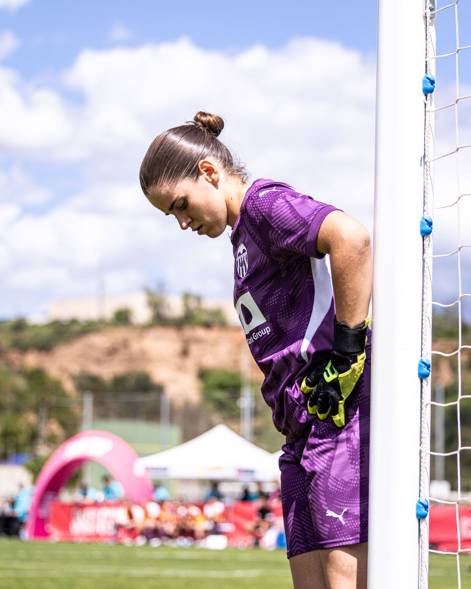 Female goalkeeper in purple kit leans against goalpost during a sunny outdoor soccer match.