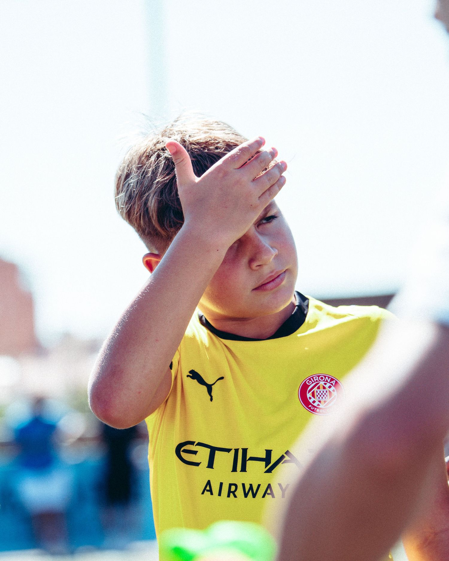 Young boy in yellow Etihad Airways Puma jersey shields his eyes from bright sunlight.