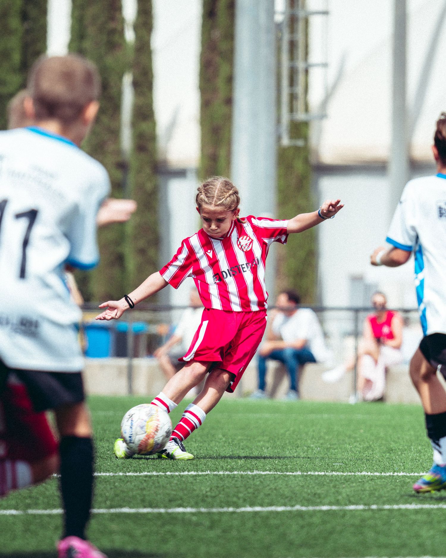 Young girl in red and white striped soccer kit taking a powerful shot during a youth match.