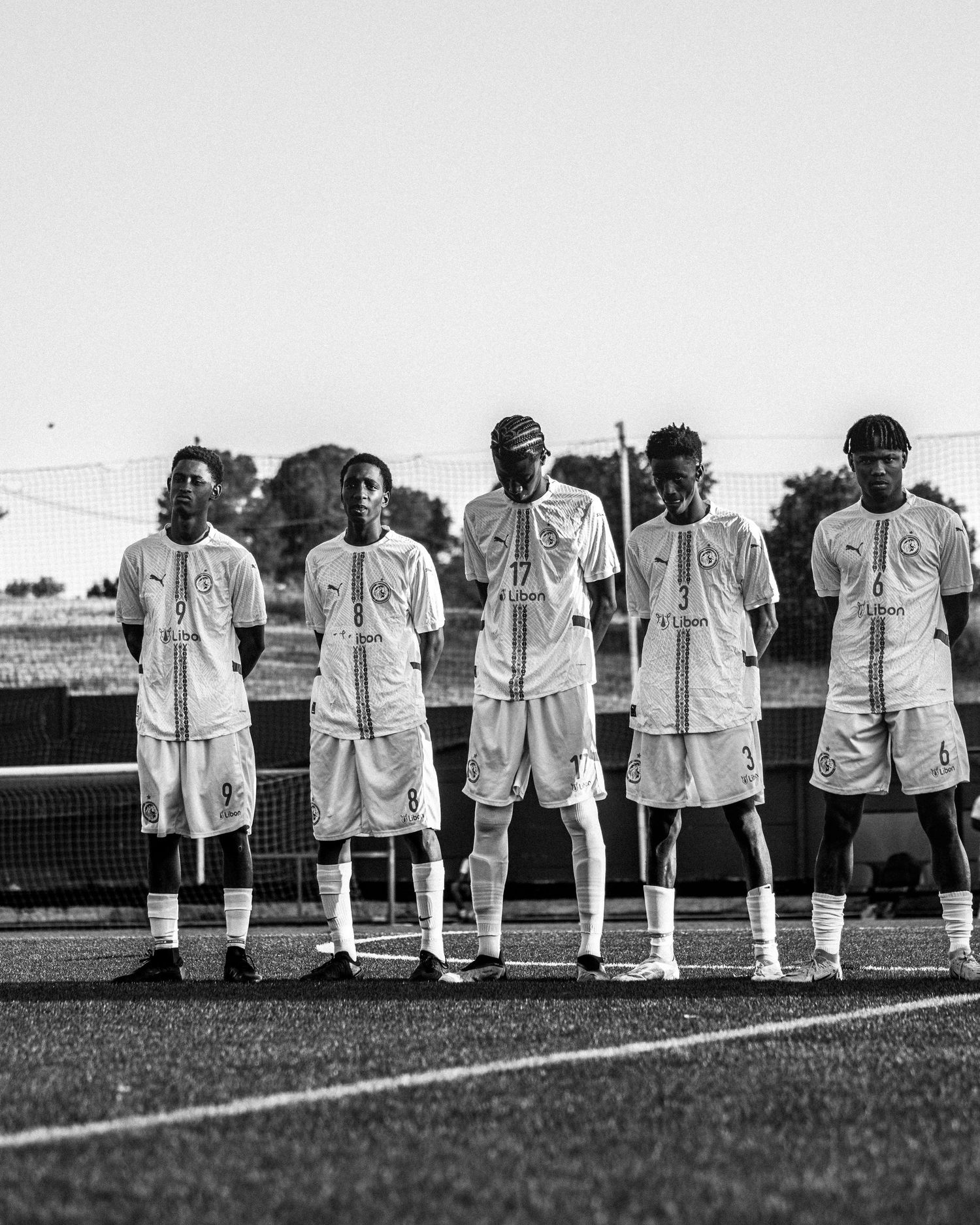 Five young soccer players in white uniforms stand in a line on a field in a black and white photo.