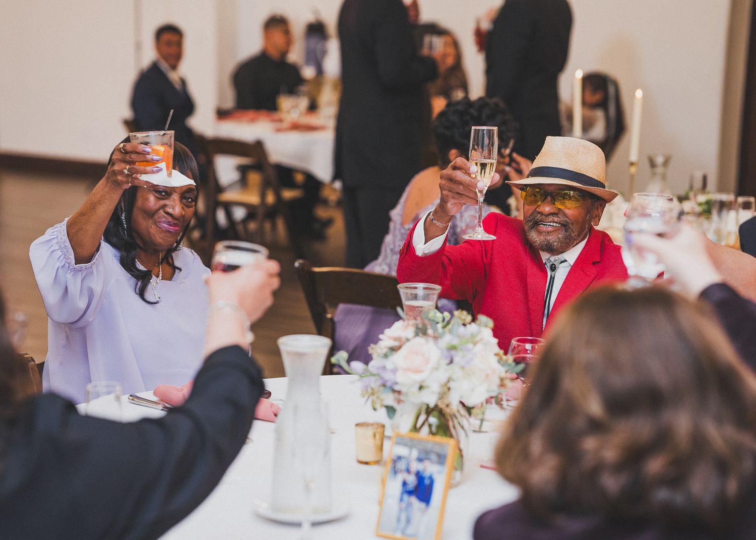 People raise their glasses in a toast at an elegant indoor celebration with white table settings and floral centerpieces.