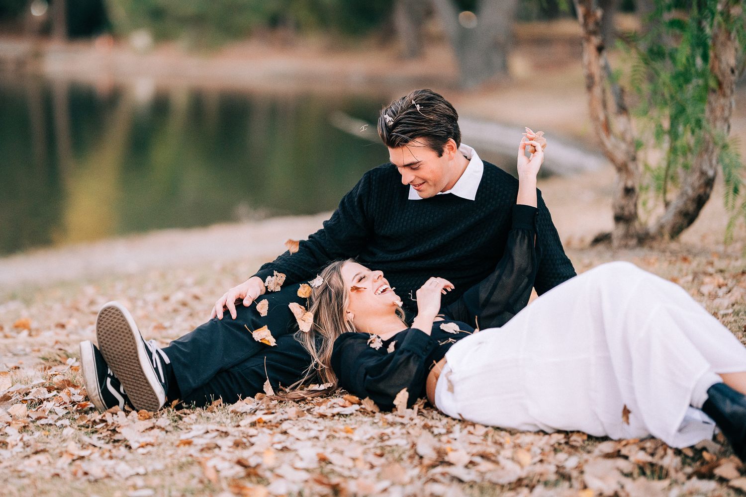 A couple laughing while sitting on fallen leaves by a lake shore in coordinated black and white attire.