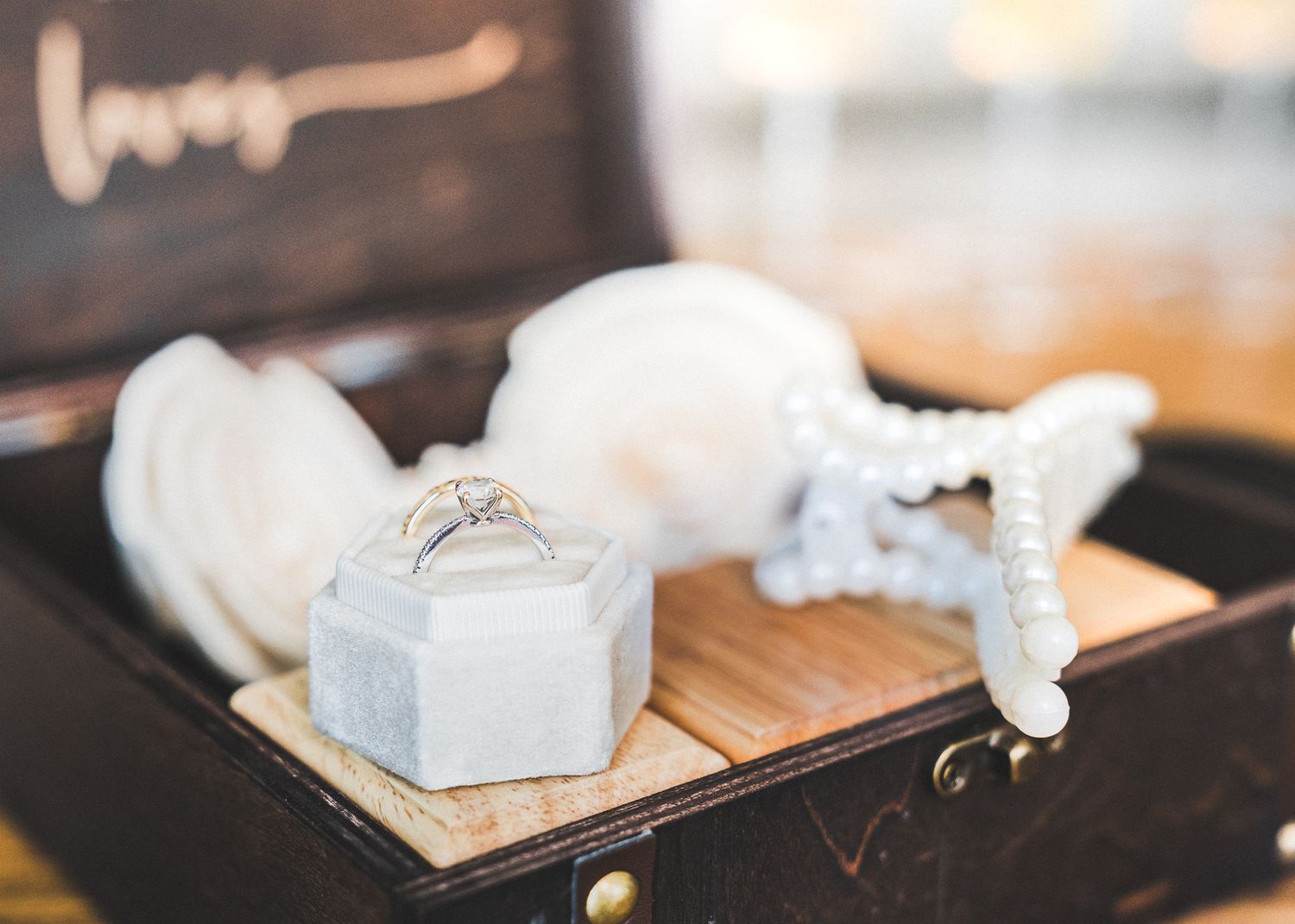 A diamond engagement ring sits in a small white box surrounded by delicate white ribbons on a wooden surface.